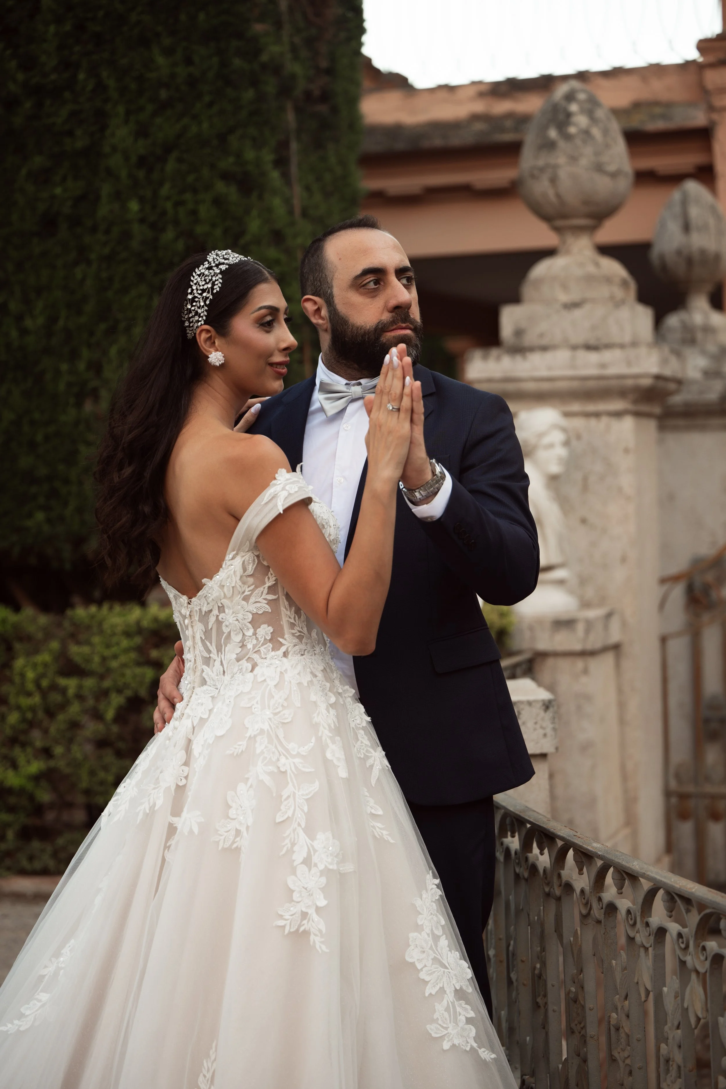 Pareja de recién casados en, vestido de novia y traje, posando en un balcón con adornos de piedra y vegetación al fondo en un día nublado.