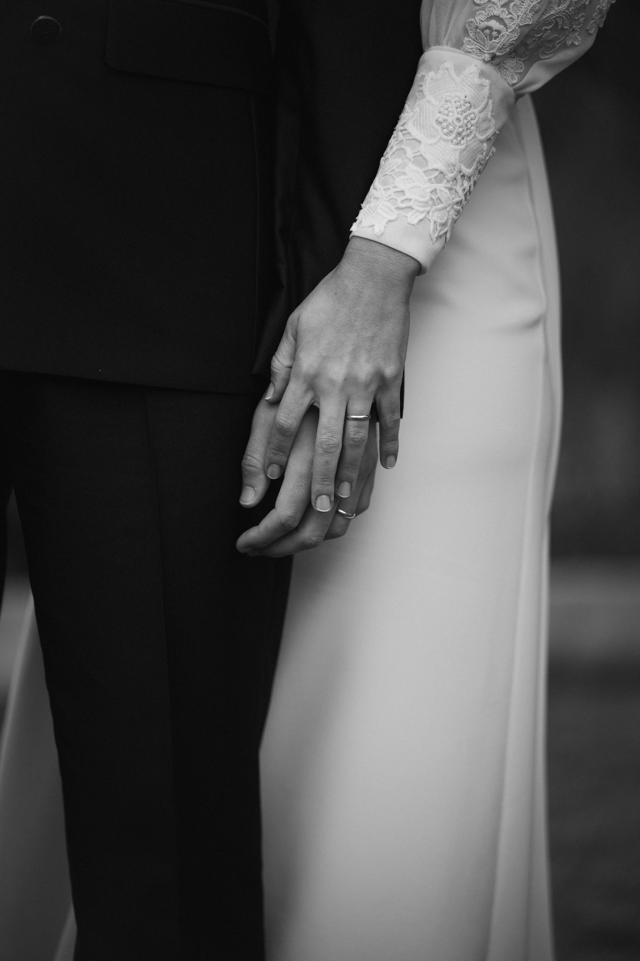Imagen en blanco y negro de una pareja, mostrando sus manos unidas con un anillo en el dedo anular de la mano izquierda, vestida con ropa formal, en lo que parece ser una escena de boda o compromiso.