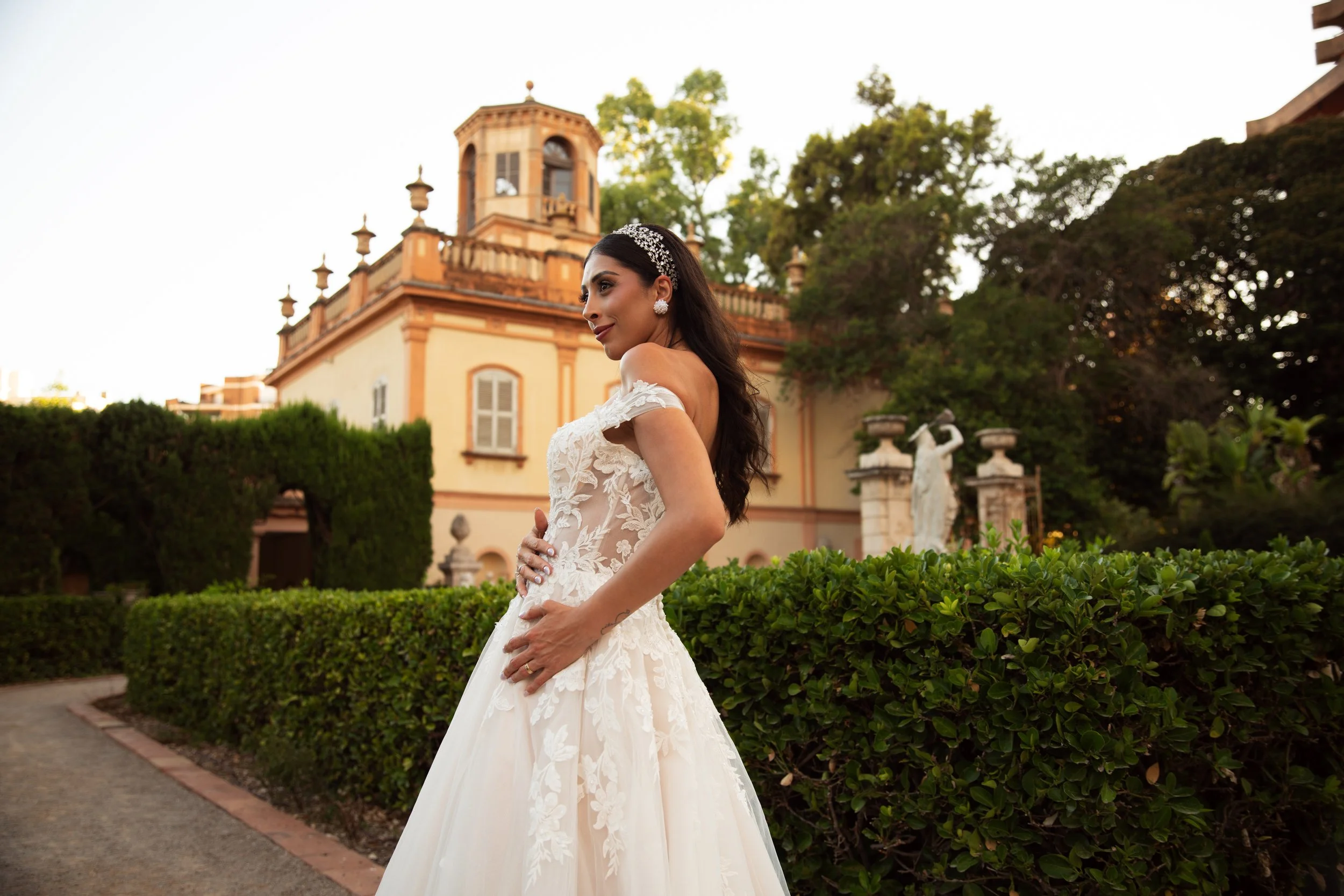 Una mujer vestida con un vestido nupcial blanco con encaje, posando frente a un edificio antiguo, rodeada de jardines y estatuas, en un entorno elegante y romántico, al atardecer.