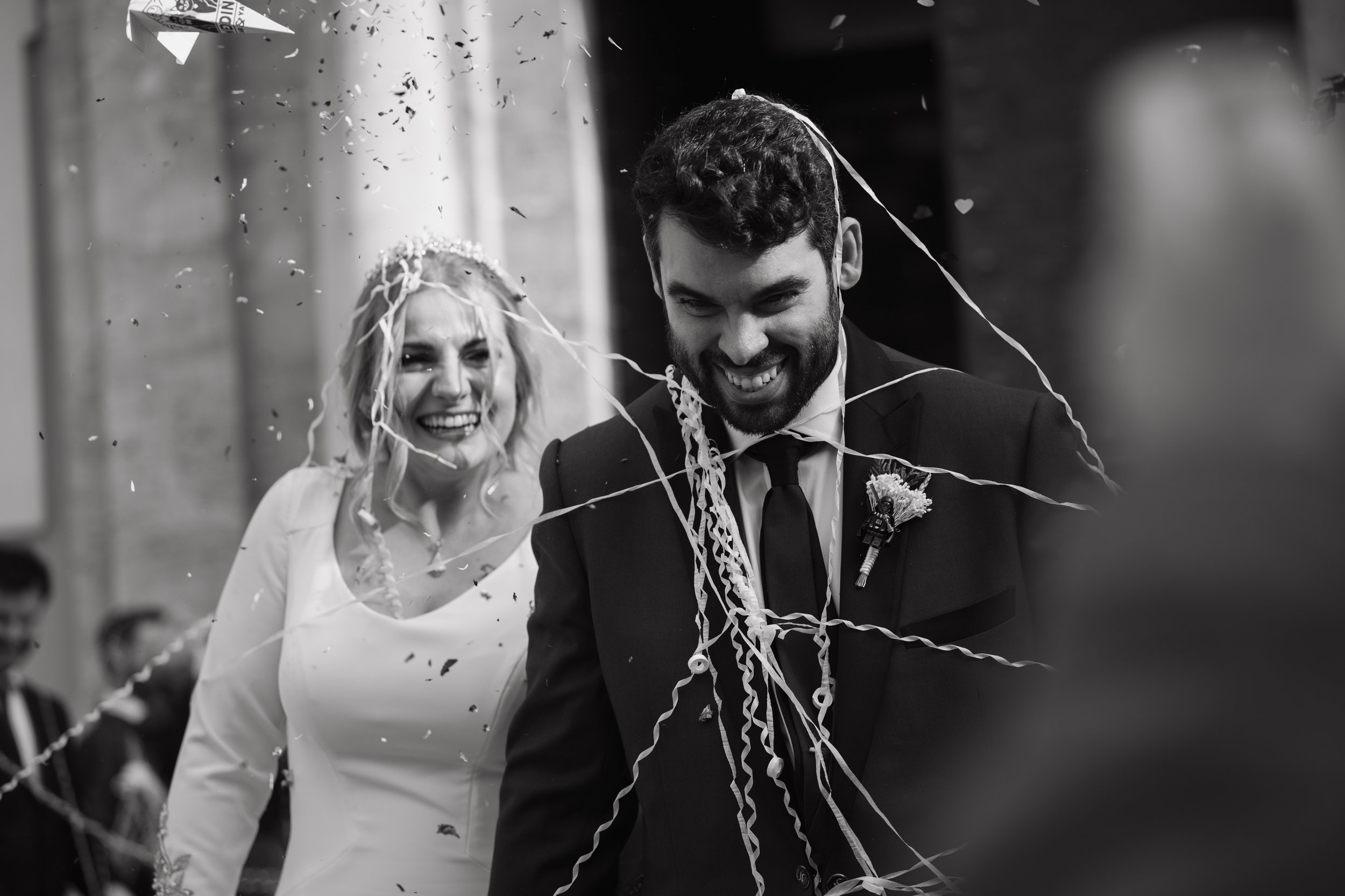 Una pareja de novios en su boda, sonriendo y cubiertos de serpentinas y confeti en un ambiente de celebración.