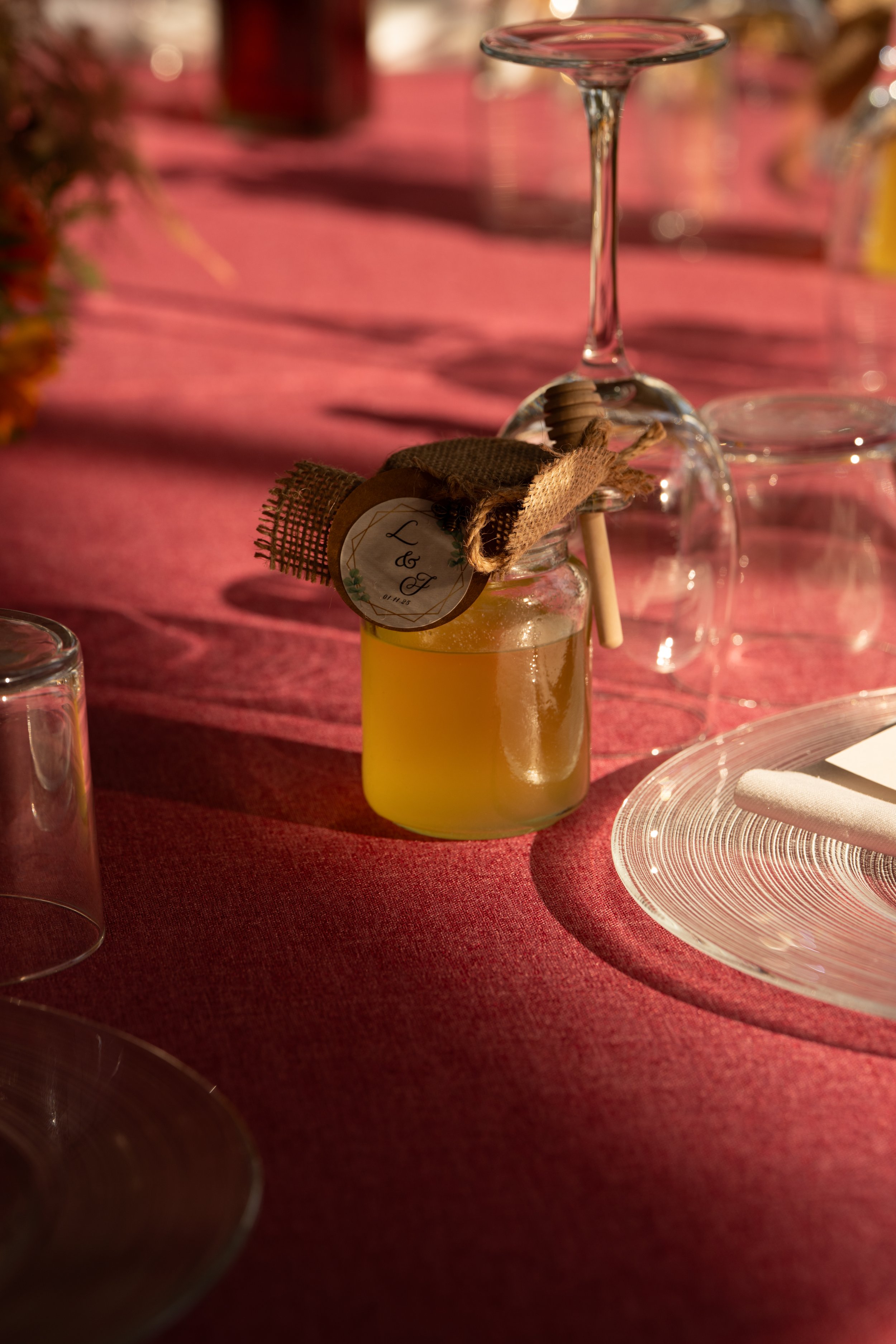 Vela amarilla en un frasco con etiqueta y decorada con cinta de arpillera, colocada sobre una mesa con mantel rojo y copas de vino volteadas al fondo.
