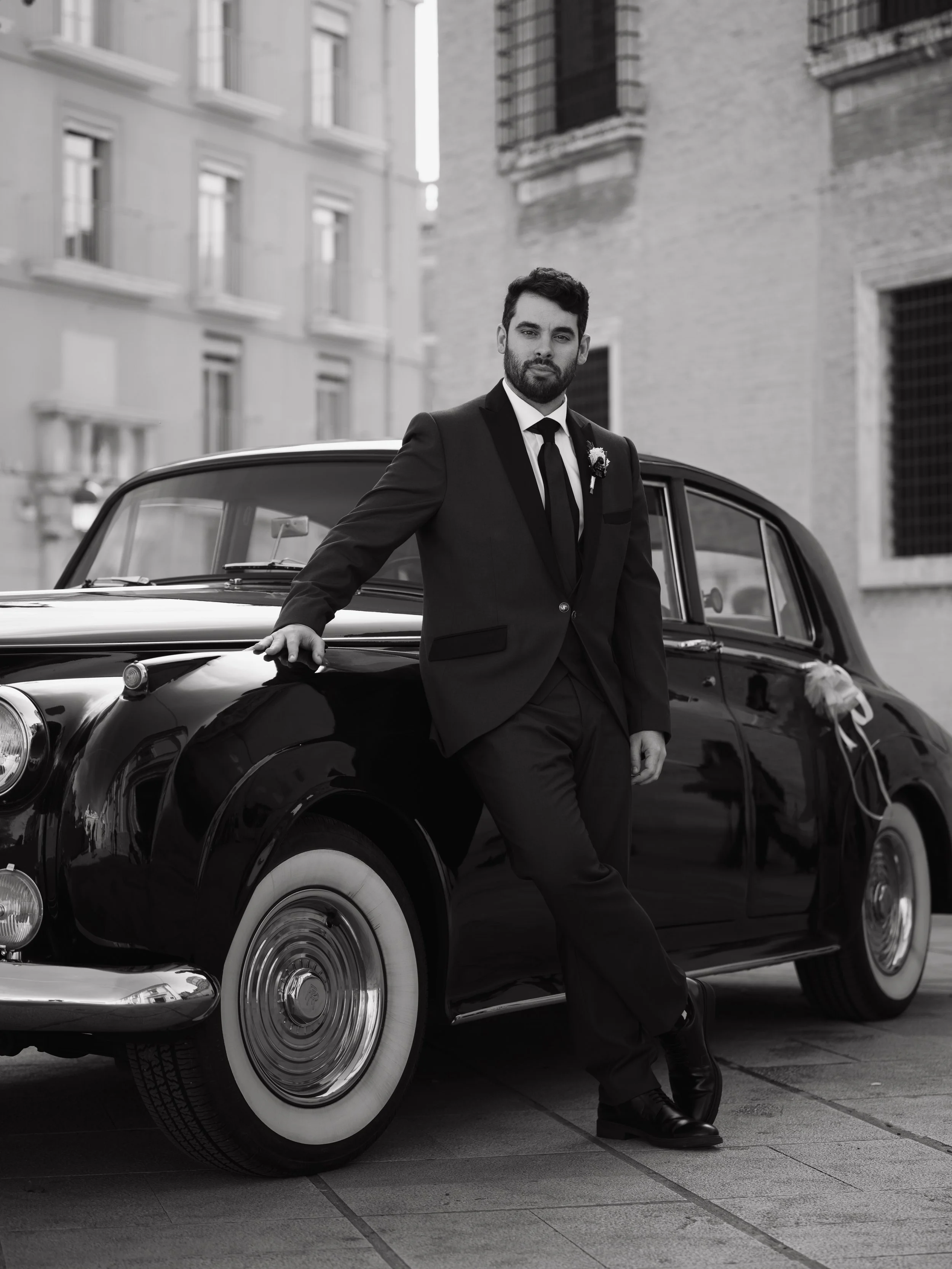 Hombre con barba y cabello oscuro, vestido con traje negro, corbata y flor en la solapa, apoyado en un automóvil clásico negro, en una calle urbana con edificios de fondo, en blanco y negro.