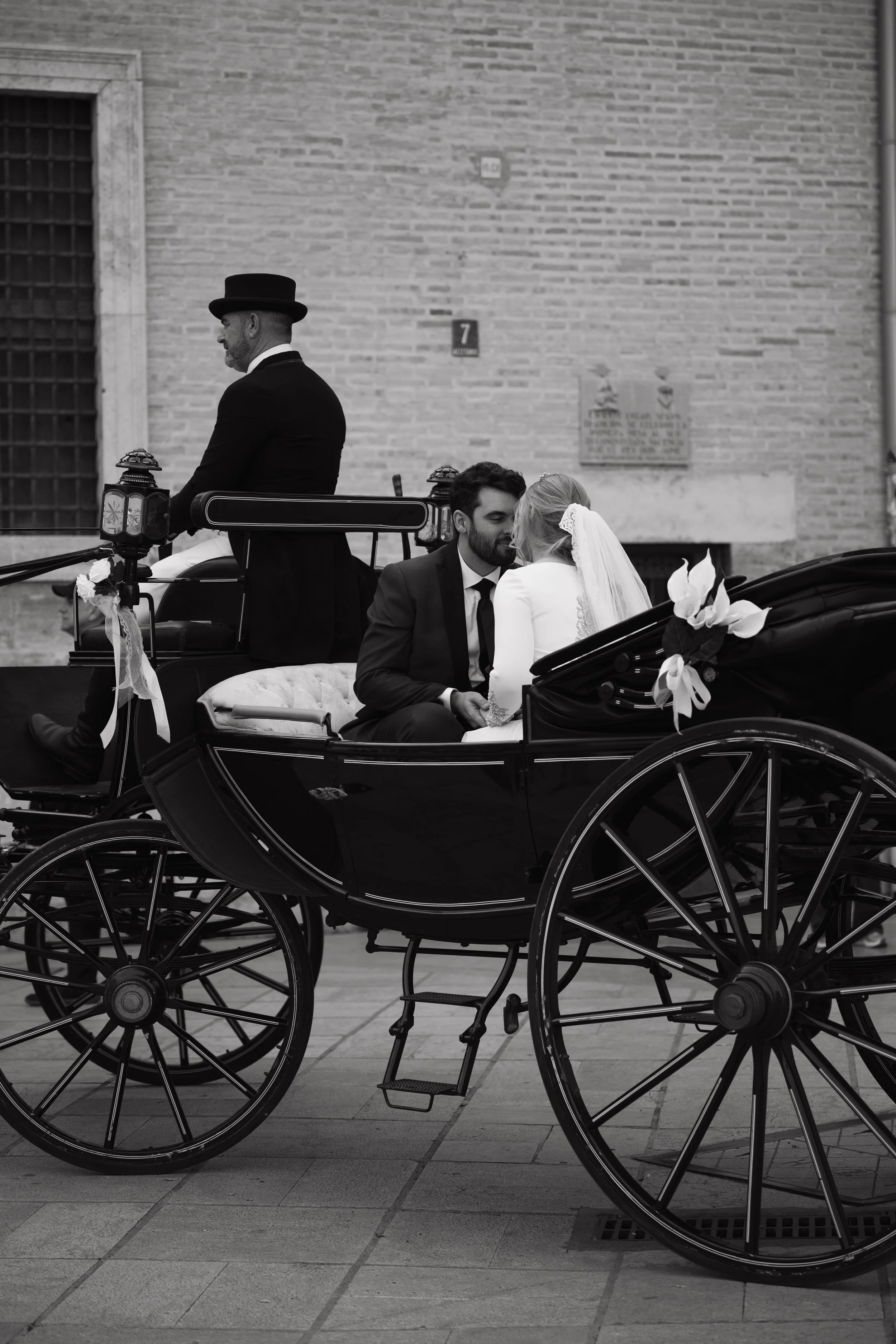 Escena en blanco y negro de un carruaje antiguo con una pareja en la parte trasera y un cochero en la parte delantera, con una pared de ladrillos en el fondo.
