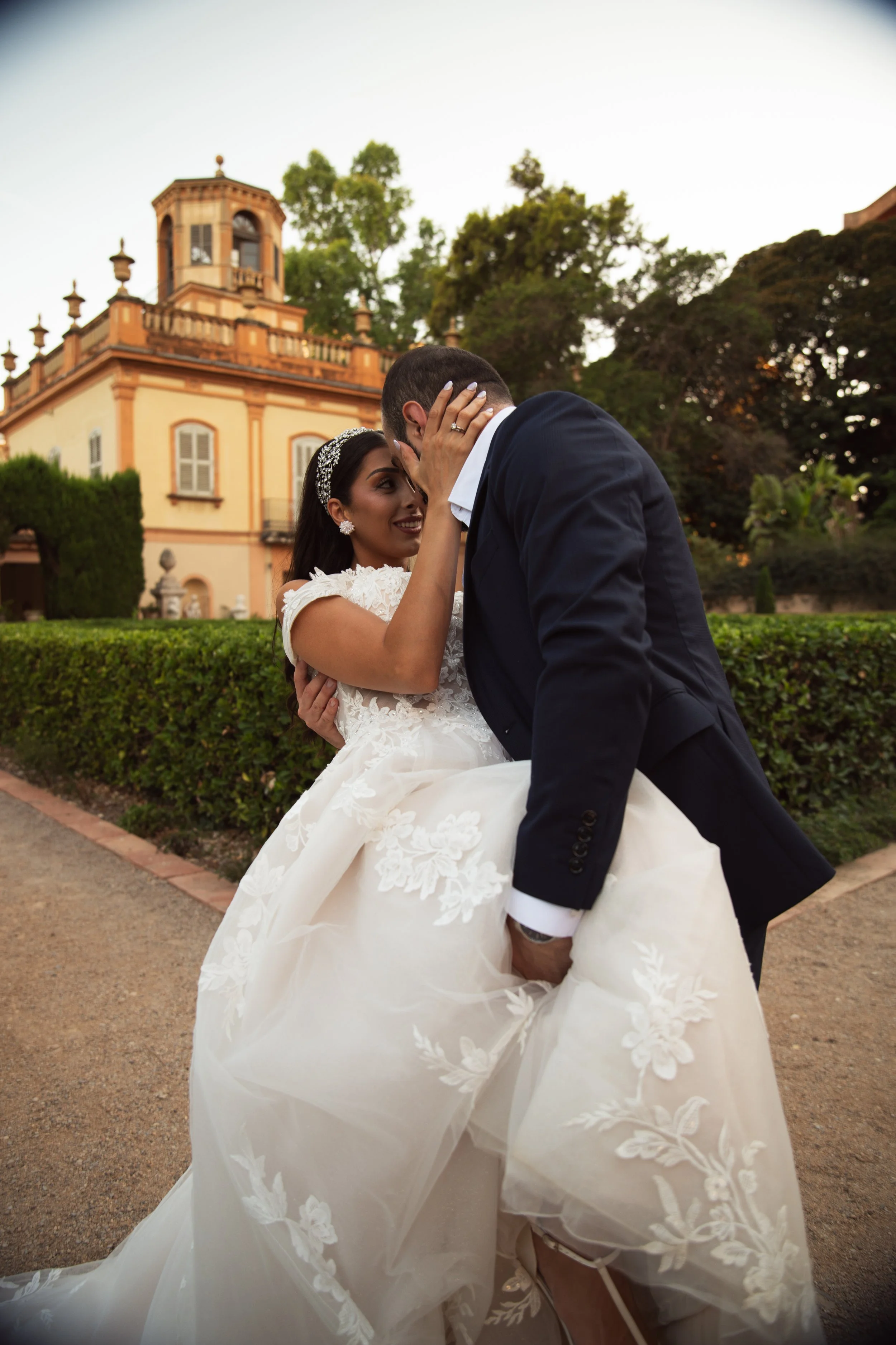 Pareja de boda en un jardín, la novia lleva vestido blanco con encaje y tiara, el novio lleva saco oscuro y camisa blanca, están en un abrazo romántico frente a una casa antigua con árboles.