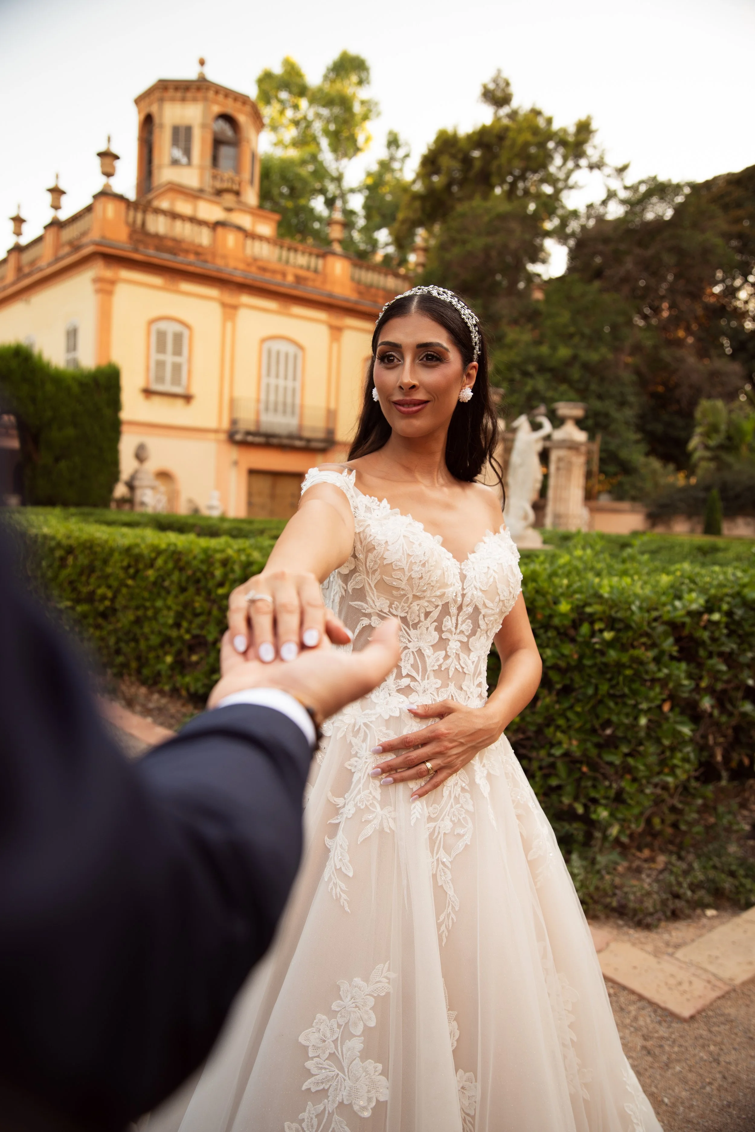 Mujer vestida de novia en una boda, extendiendo la mano para un apretón, con un fondo de árboles y un edificio histórico.