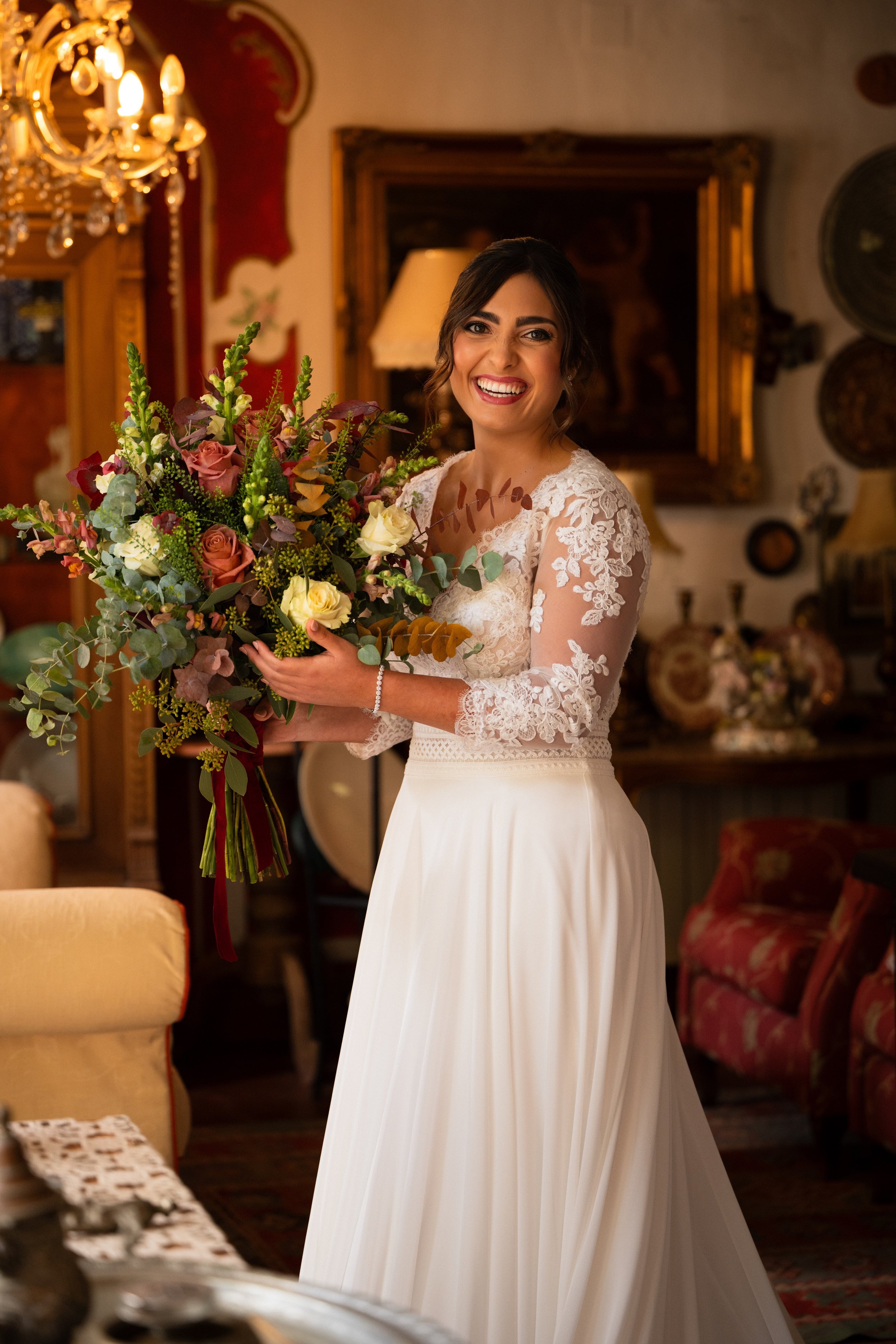 Mujer sonriendo con vestido blanco y cabello oscuro, sosteniendo un ramo de flores en una habitación decorada con muebles y cuadros antiguos.