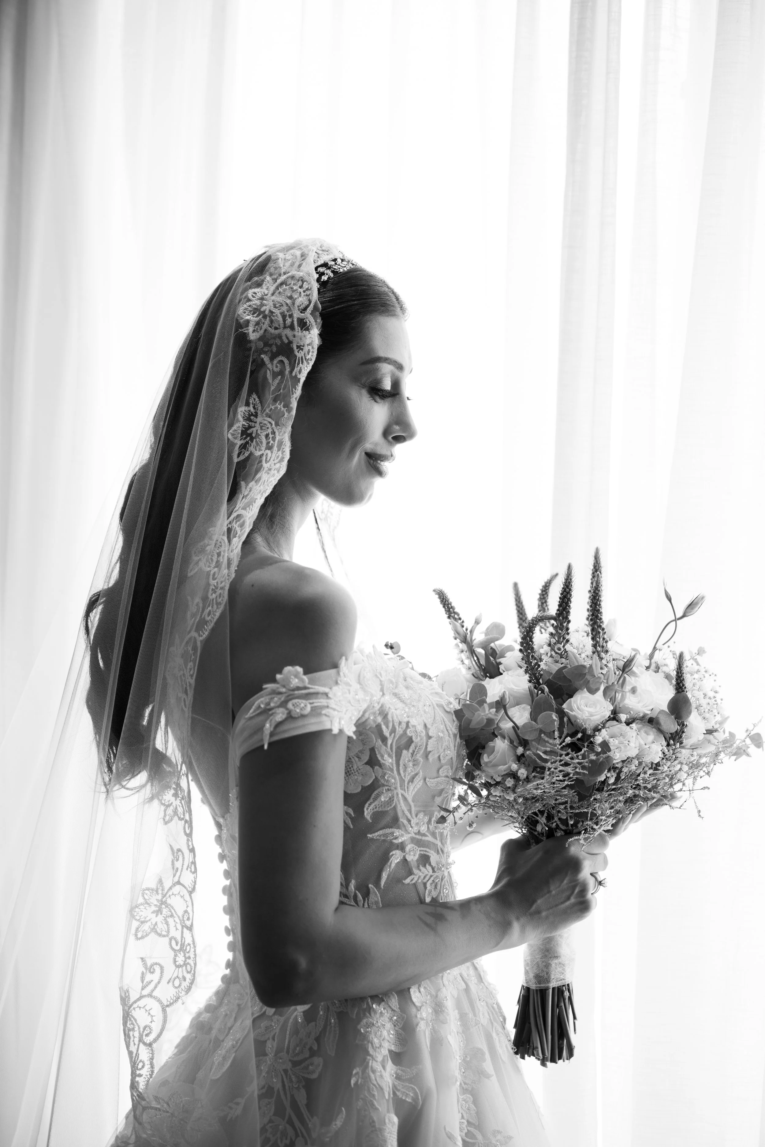 Mujer con vestido de novia y velo, sosteniendo un ramo de flores en un fondo de cortinas blancas.