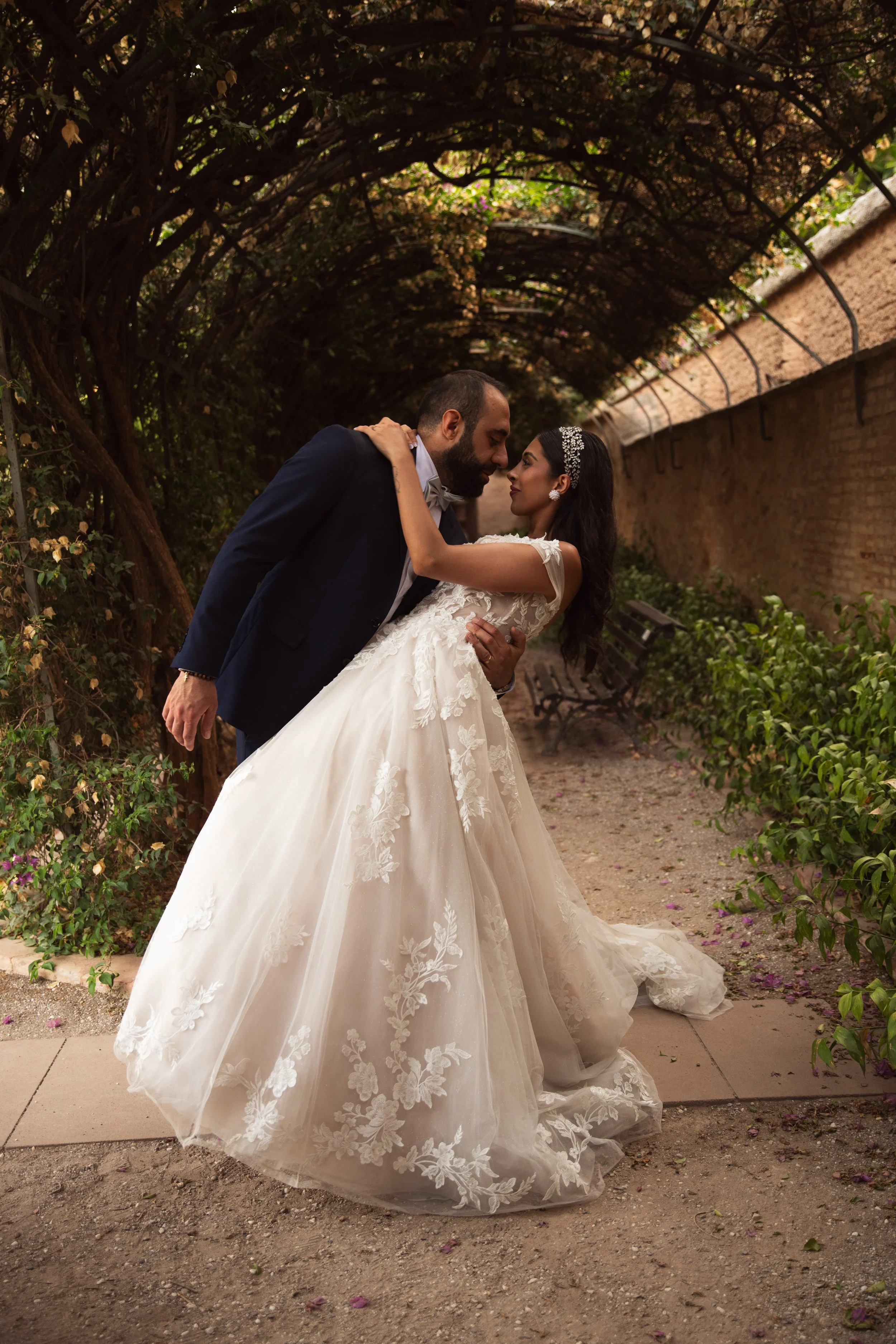 Pareja de novios en vestido de novia y traje de novio en un pasillo cubierto de enredaderas y plantas, en un ambiente romántico y natural.