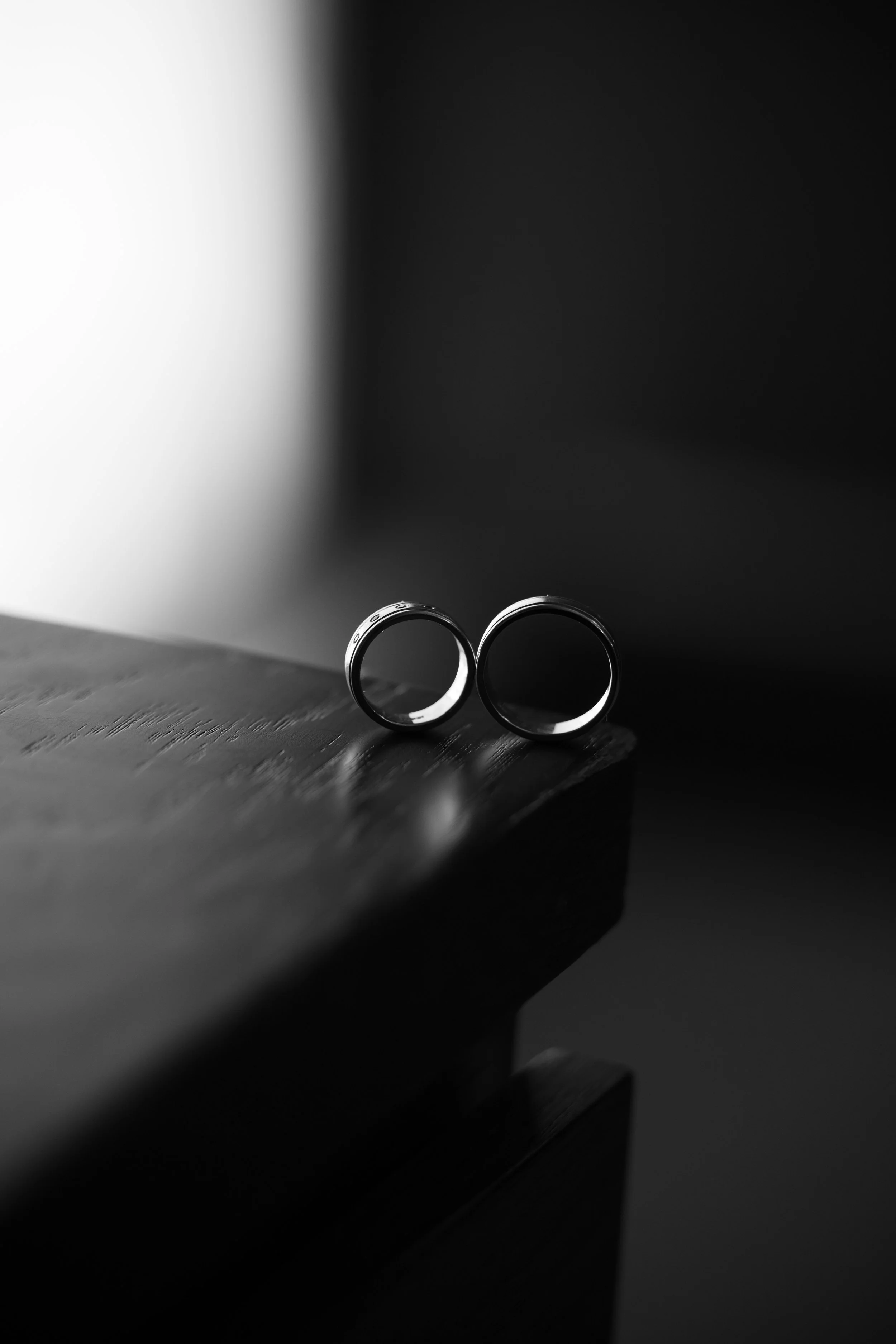 Dos anillos de boda plateados sobre una mesa de madera, con sombra y fondo oscuro.