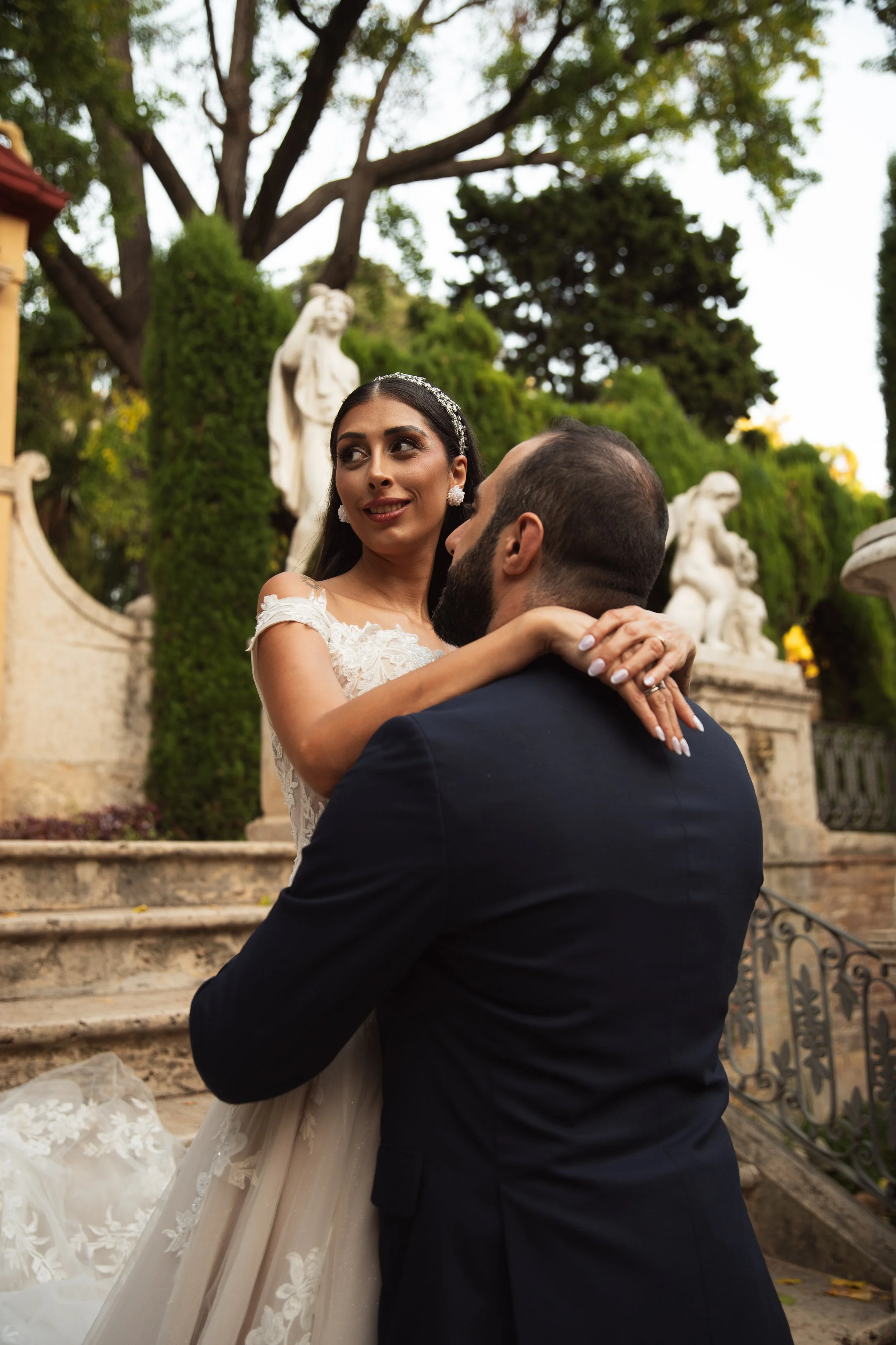 Pareja bailando en un jardín con estatuas y árboles, ella lleva vestido de novia y él traje formal.
