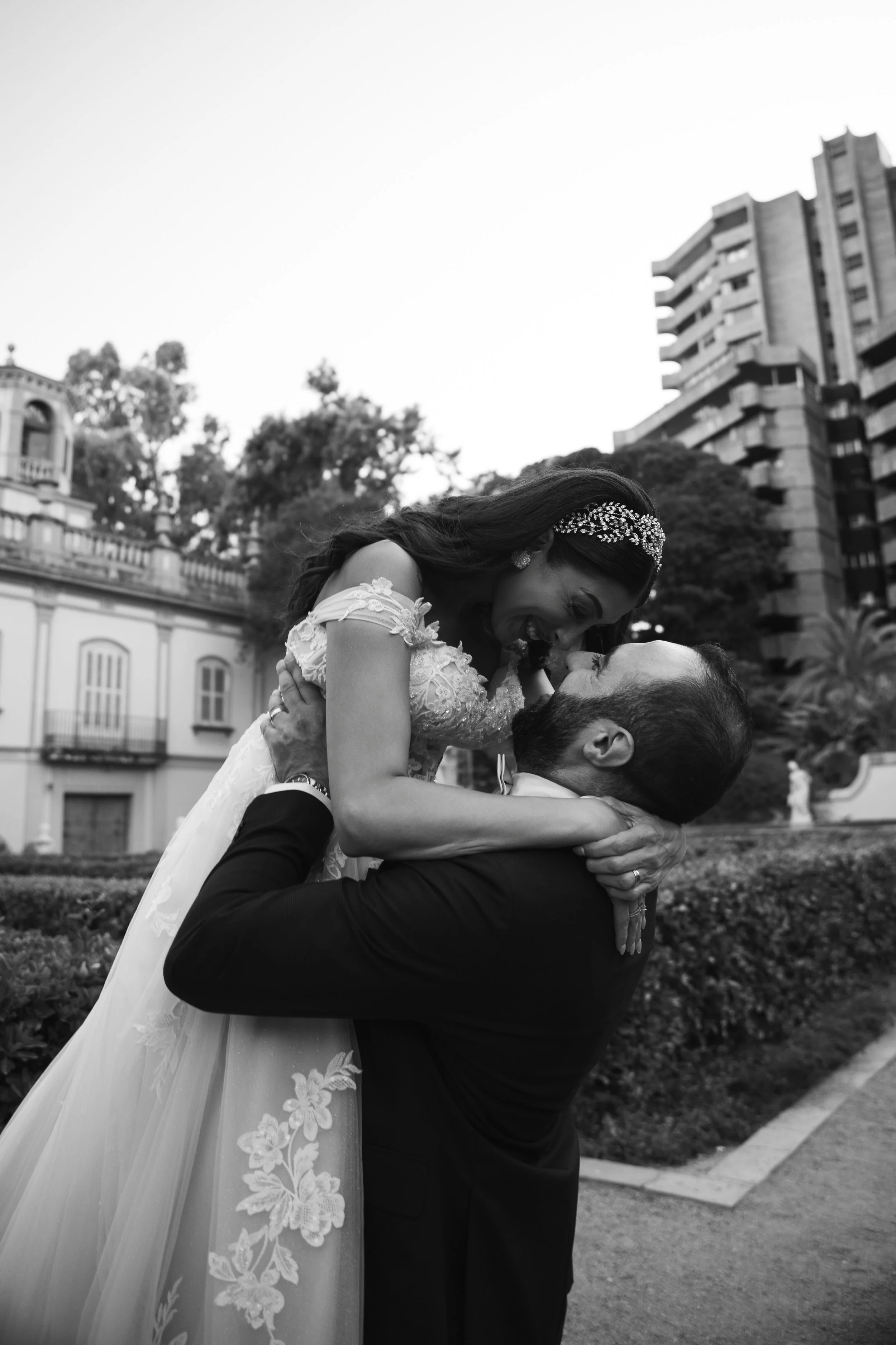 Pareja de recién casados en un abrazo fuerte en un parque, ella usando un vestido blanco con encaje y él con traje negro, fondo de árboles y edificio