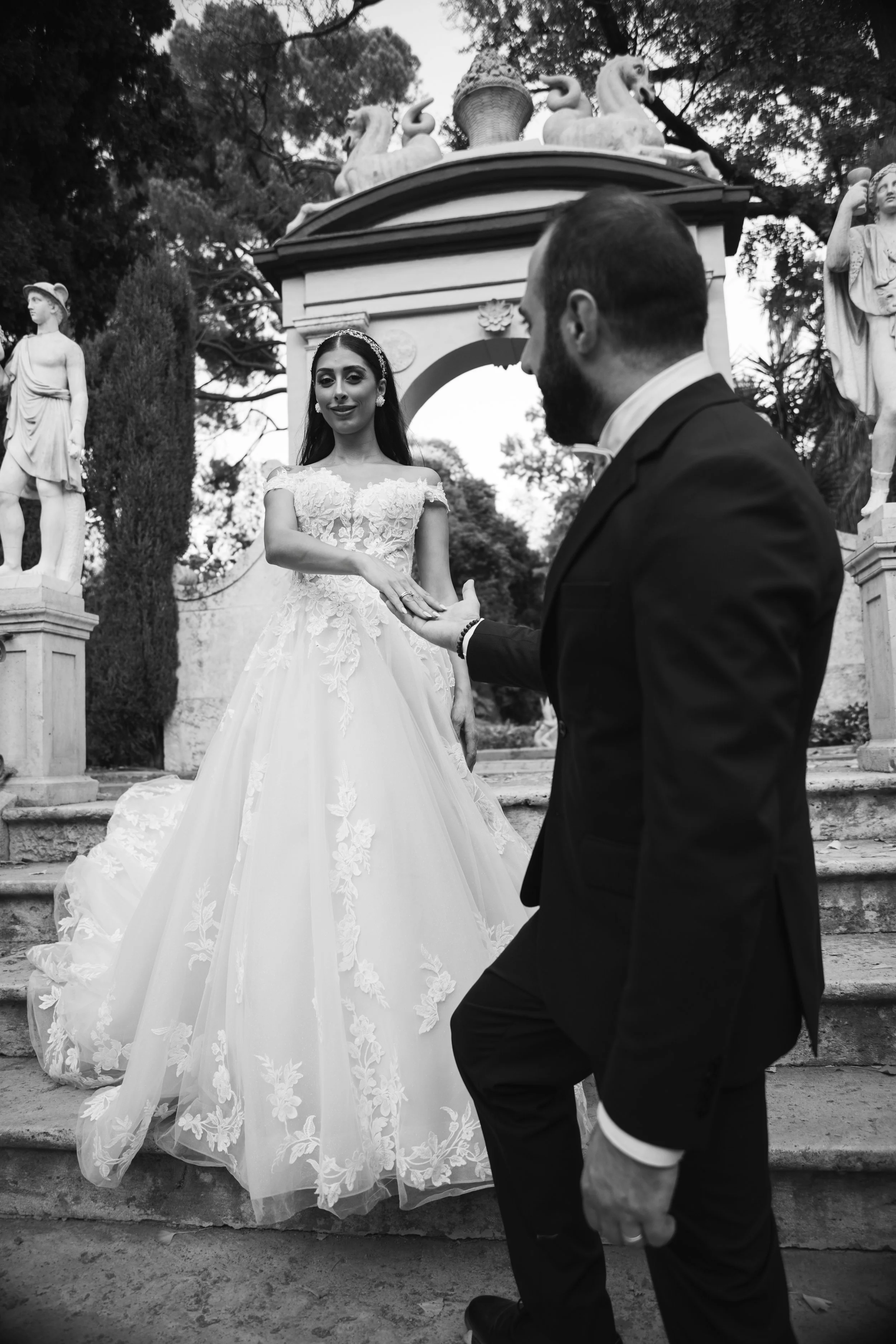 Una pareja en una boda sosteniendo las manos durante una ceremonia en un lugar al aire libre con estatuas y árboles en el fondo, en blanco y negro.