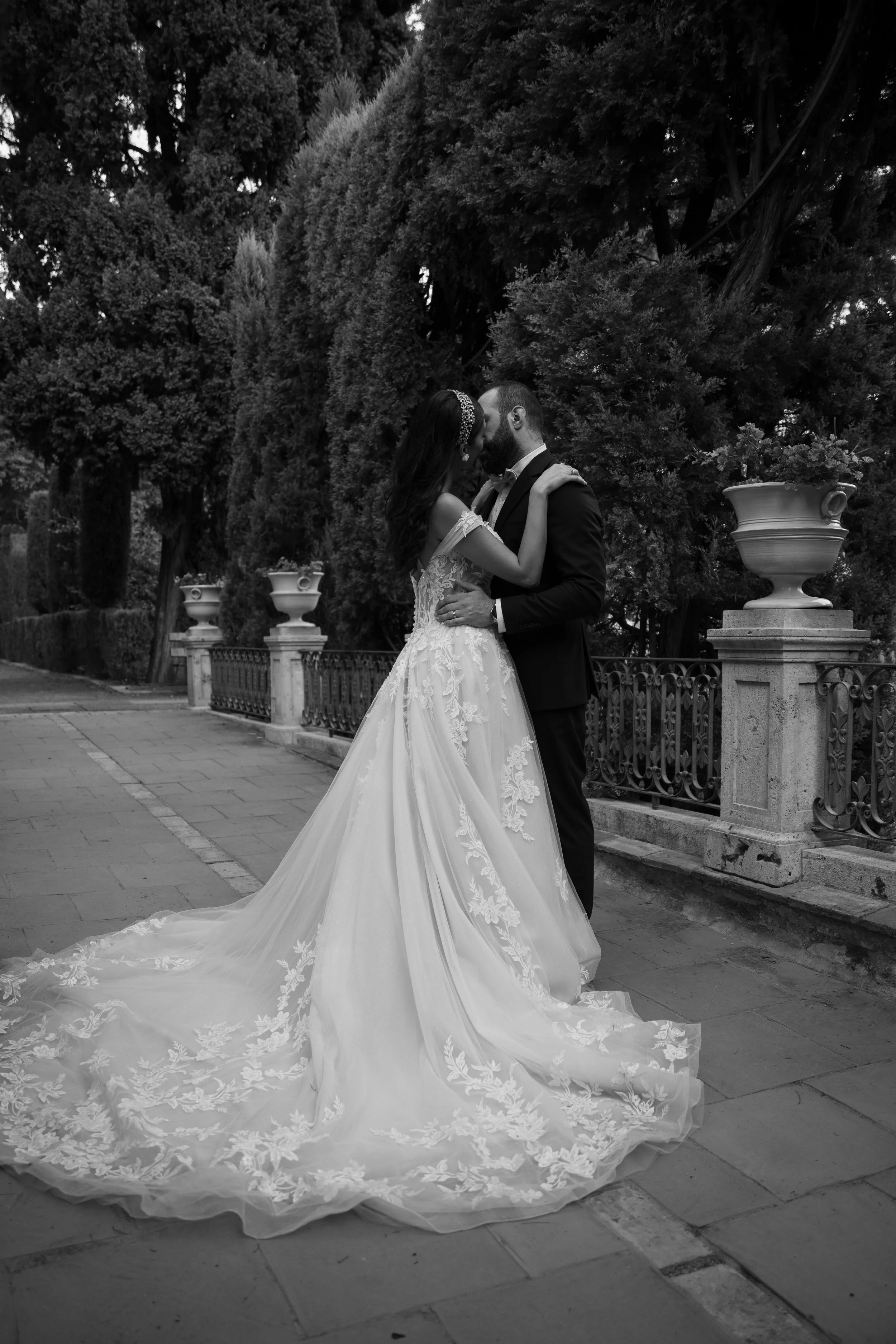 Pareja de novios en boda, la mujer con vestido largo y el hombre con traje, en un parque con árboles y macetas decorativas.