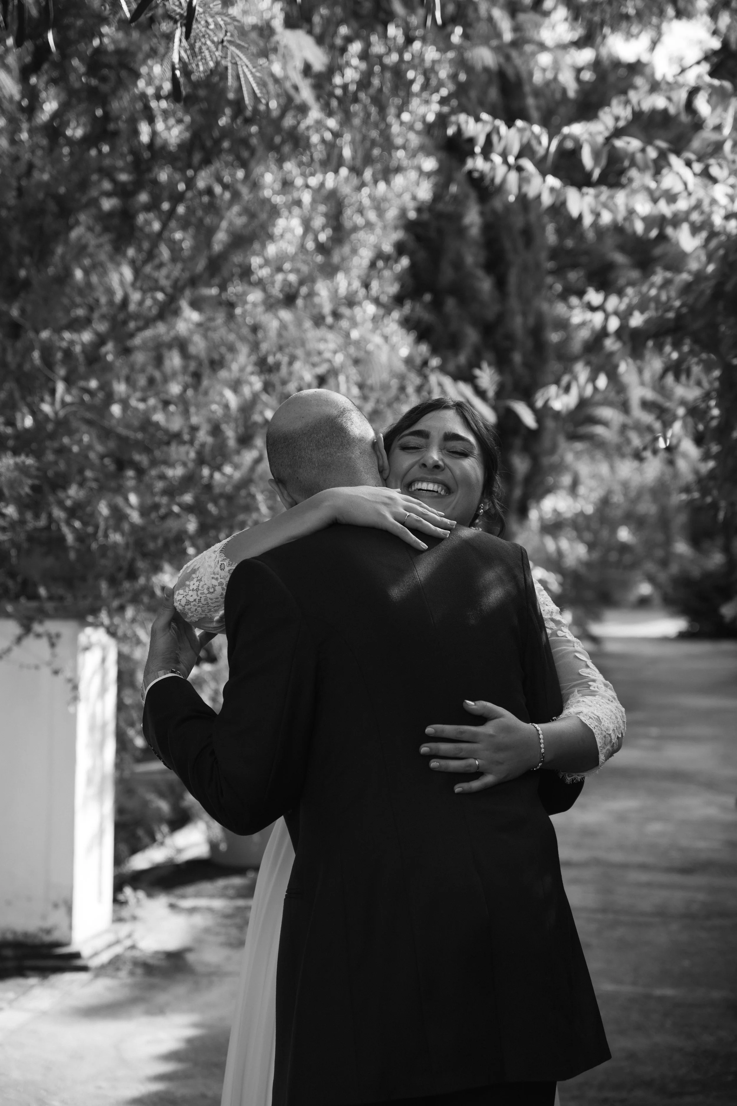 Pareja abrazándose en un parque, mujer sonriendo con los ojos cerrados y hombre de espaldas, en un entorno natural con árboles.