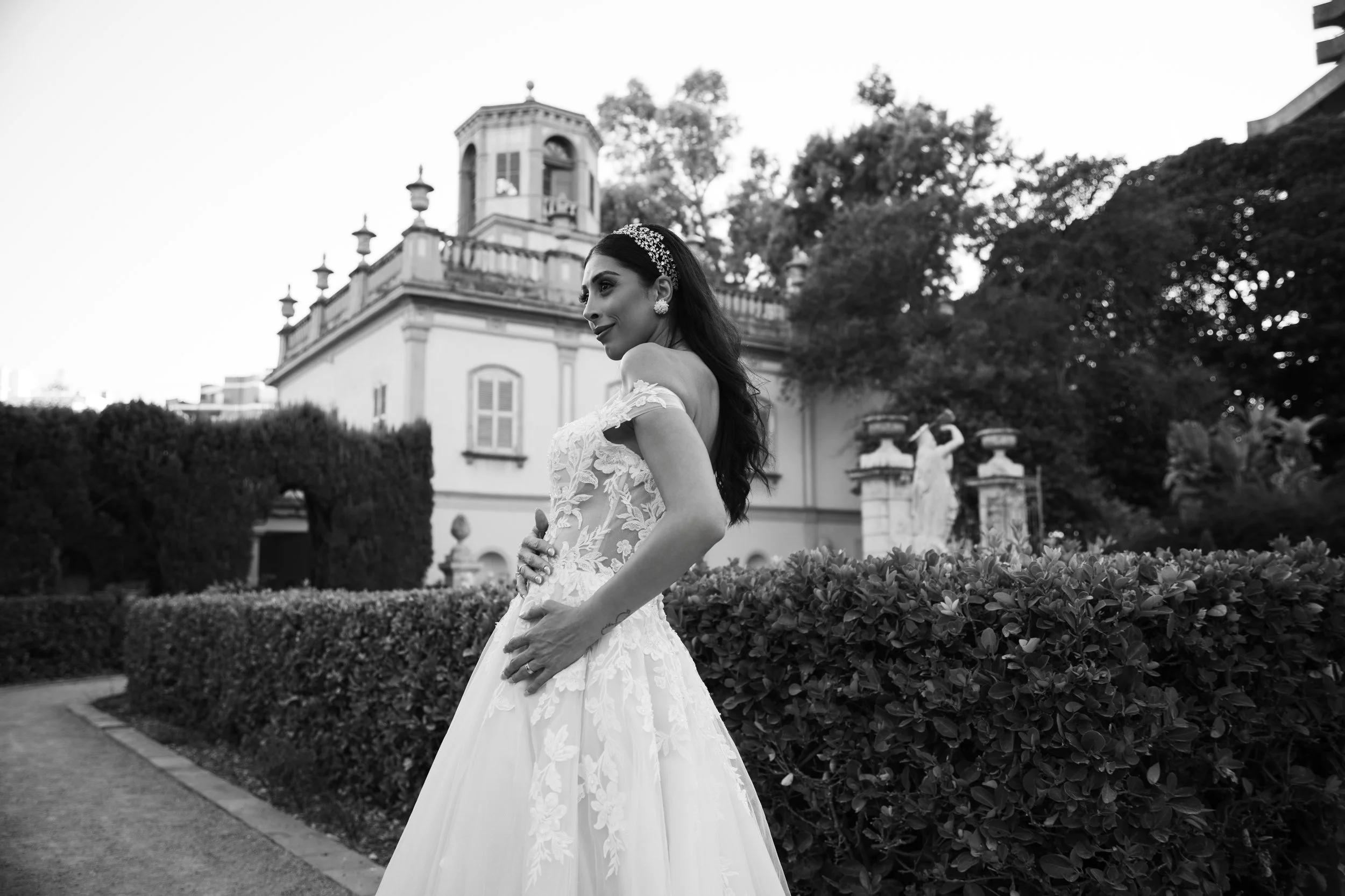 Mujer vestida con un vestido de novia en un jardín con árboles y una edificación elegante en el fondo, en blanco y negro.