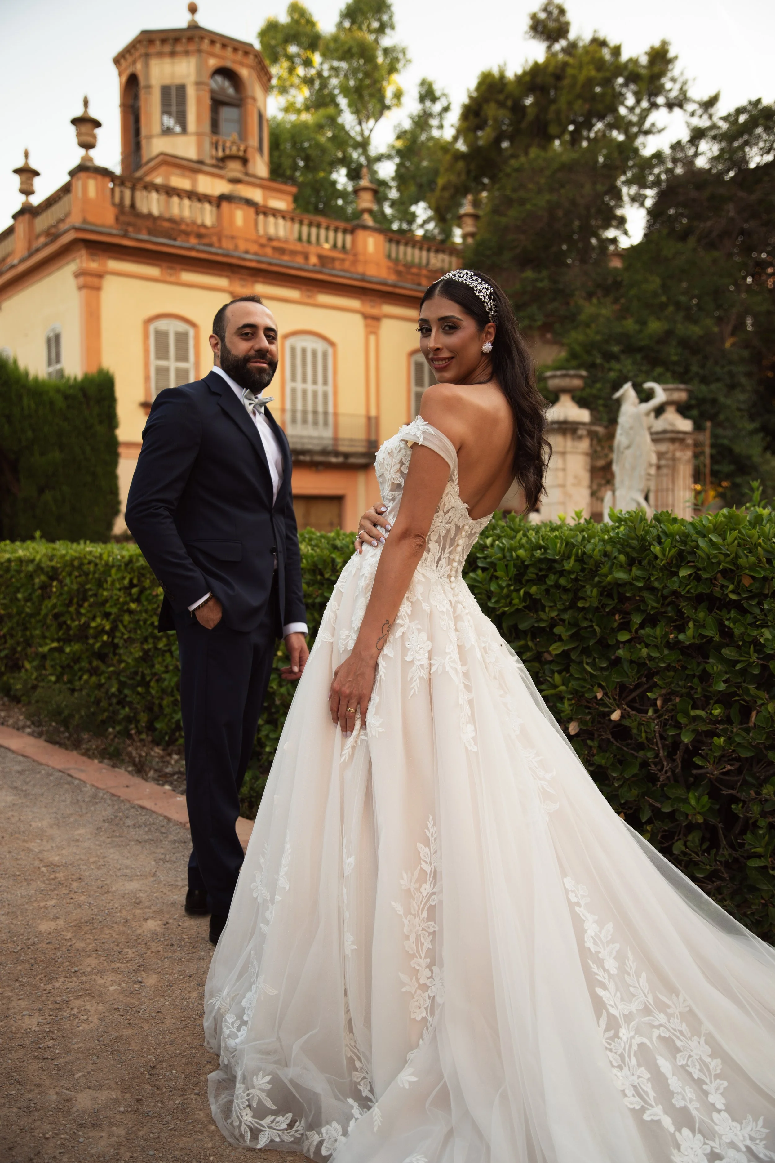 Pareja de novios en un jardín, con un edificio antiguo y estatuas en el fondo, la mujer lleva vestido de novia y el hombre traje formal.