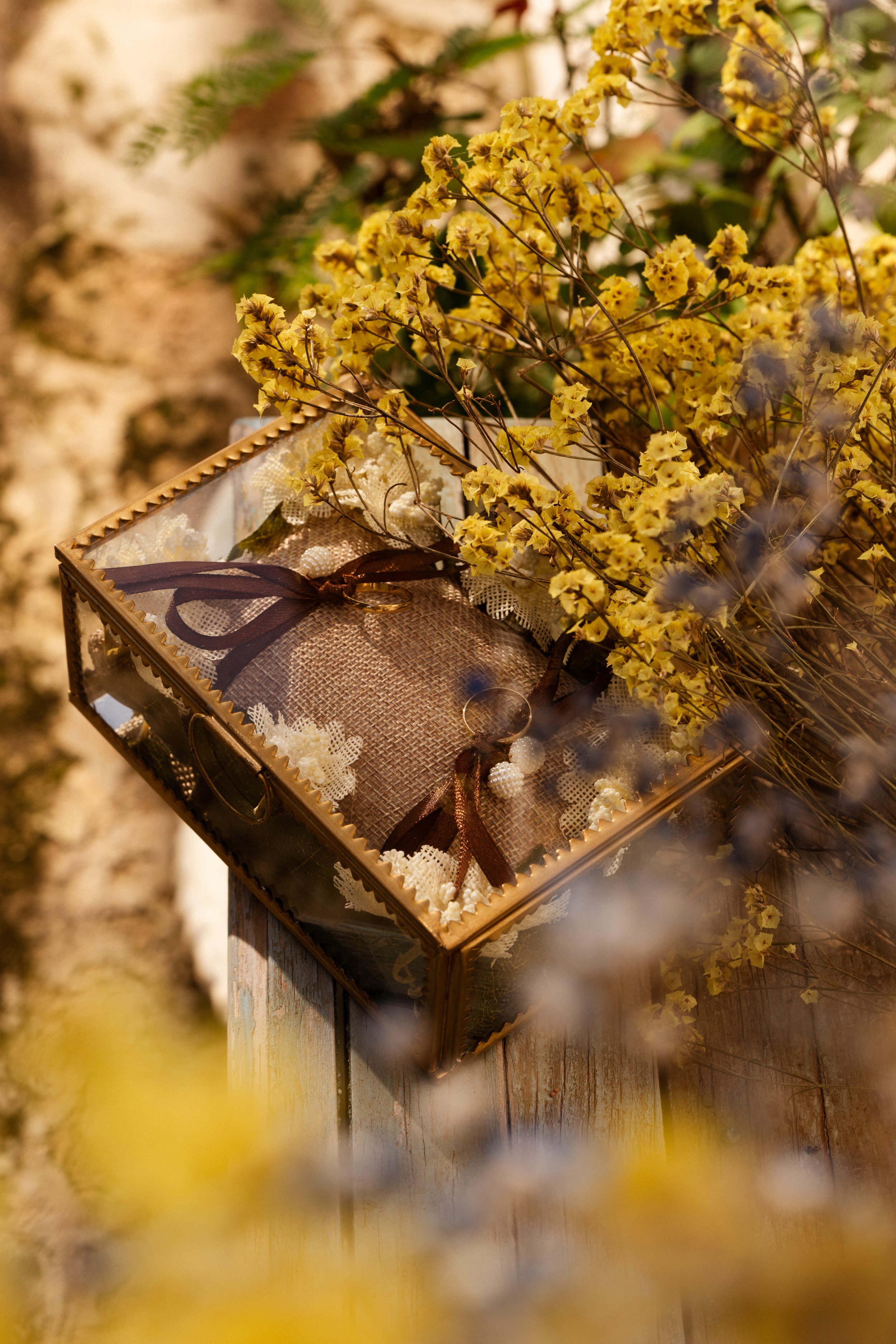 Una caja decorativa con un lazo marrón, rodeada de flores amarillas secas, sobre una mesa de madera envejecida.