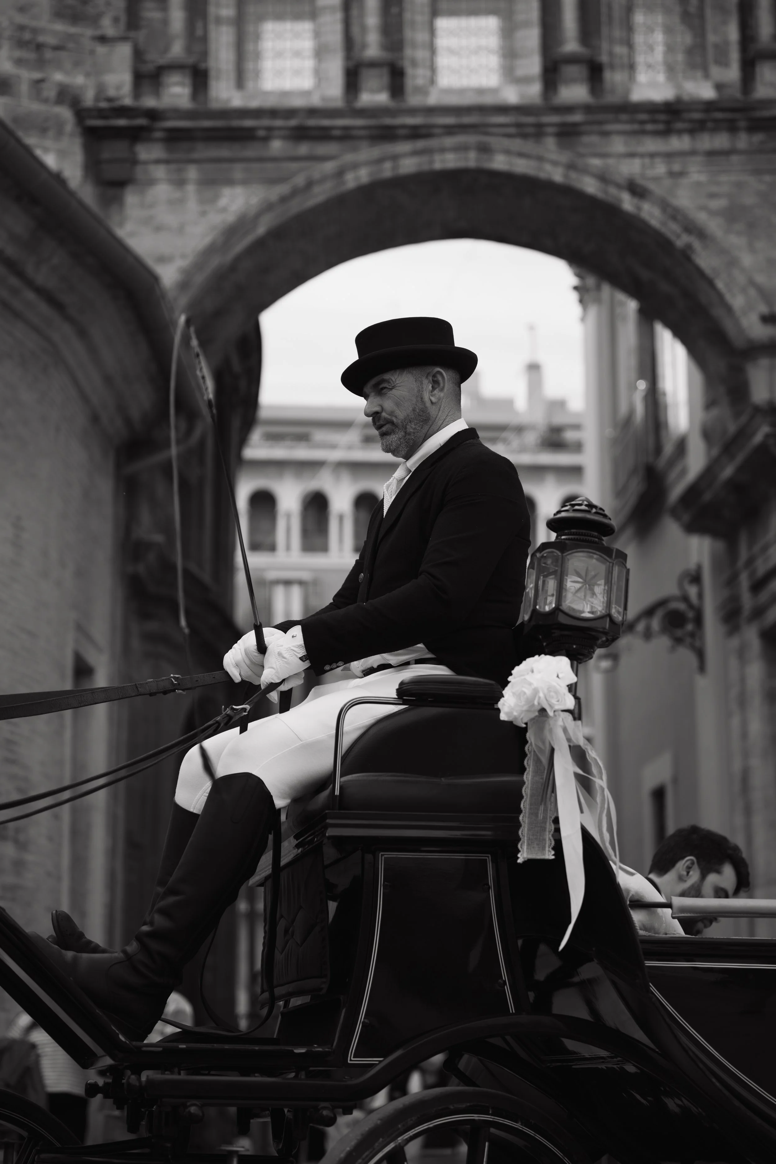 Un carruaje con un hombre vestido formalmente en su interior, usando un sombrero y guantes blancos, con una rosa en la parte trasera del carruaje, en un entorno urbano con edificios de fondo.