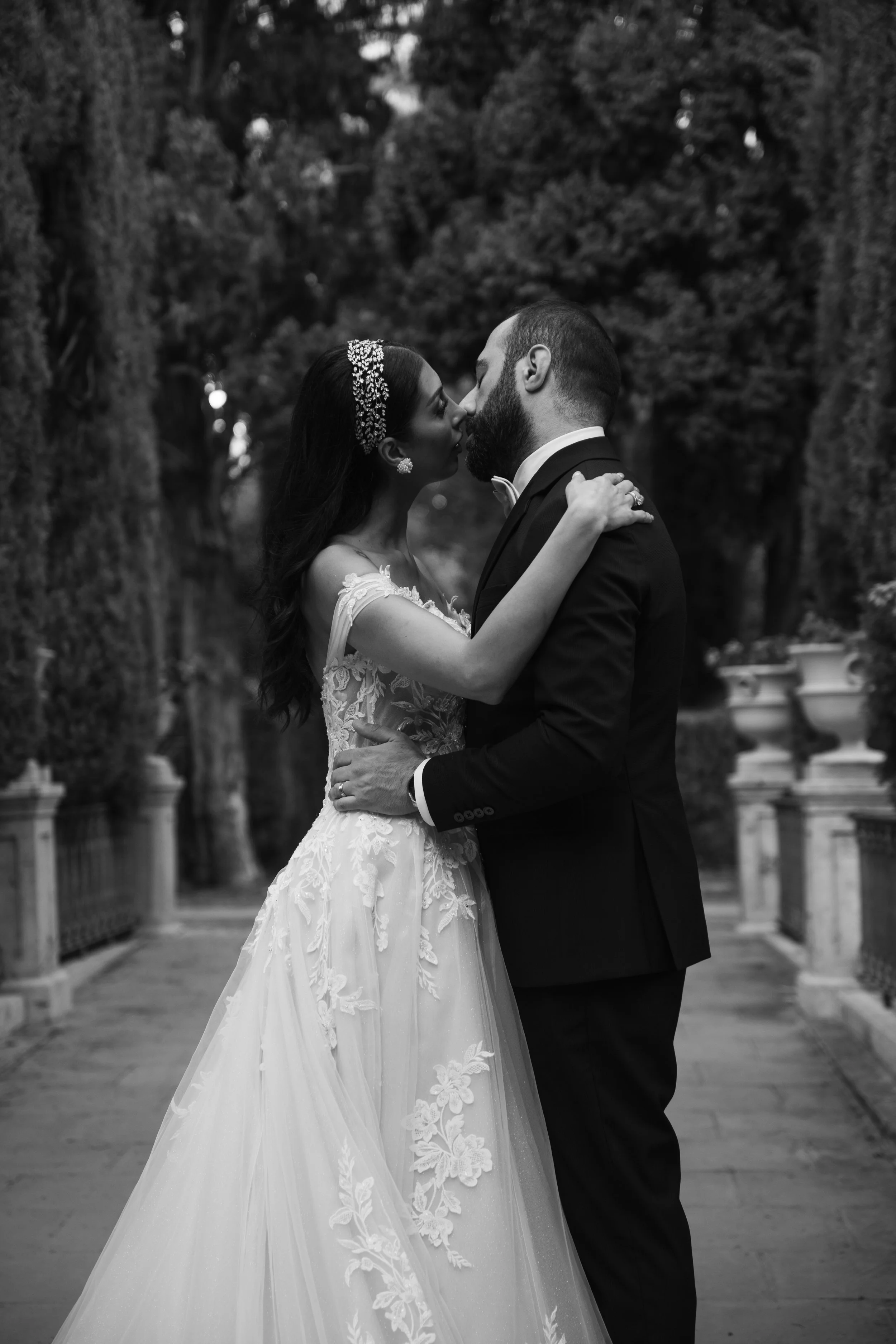 Pareja de novios abrazados, la mujer lleva vestido de novia y accesorios brillantes, el hombre con traje formal en un pasillo de parque