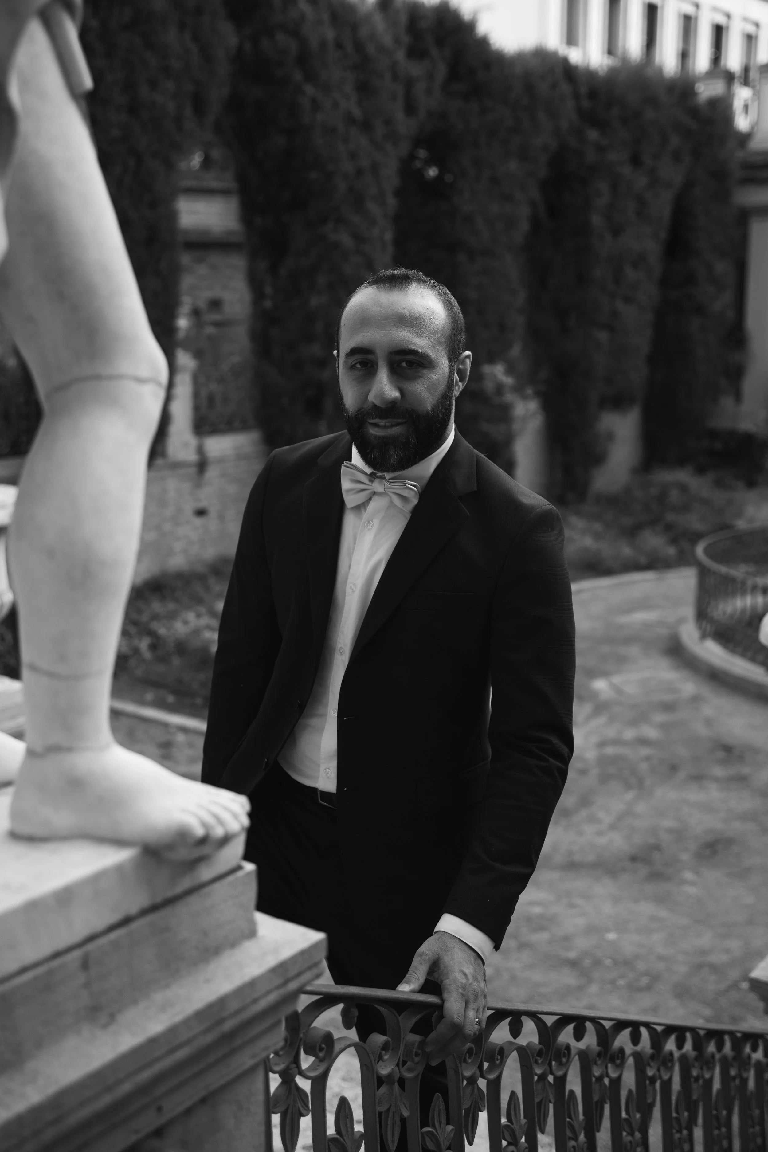 Hombre con barba y vestido formal, de espaldas, en un entorno exterior con árboles y un edificio, mirando por una cerca de hierro, en una imagen en blanco y negro