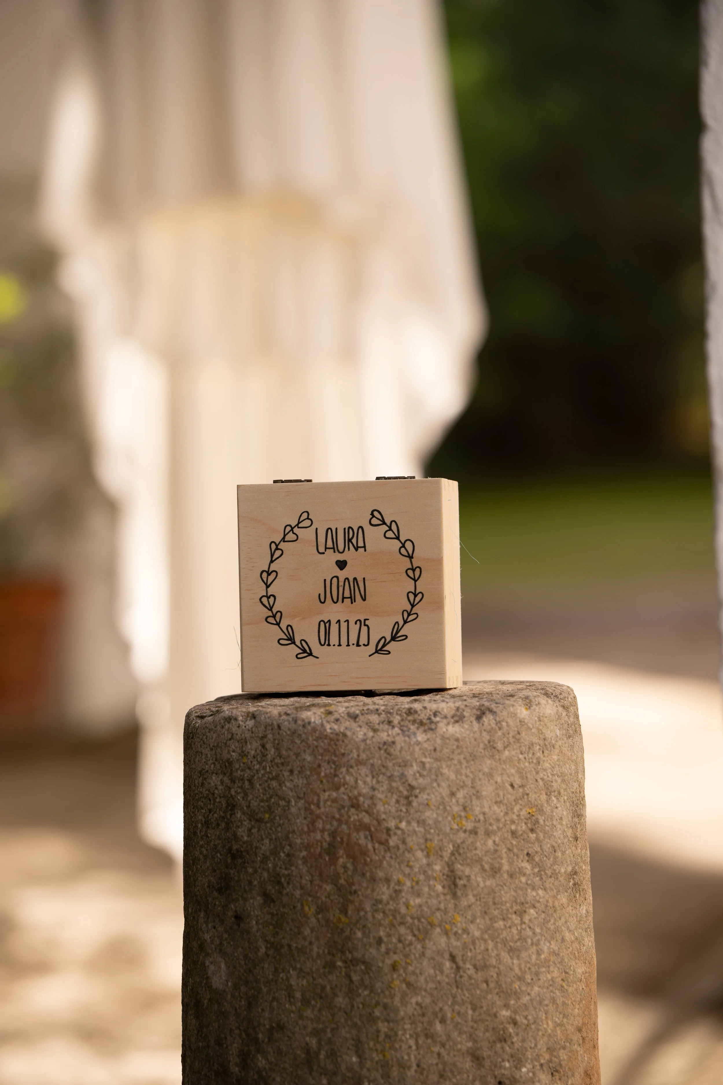 Detalle de una caja de madera con nombres y fecha, ubicada sobre una piedra, al aire libre con fondo difuso de plantas y estructura.