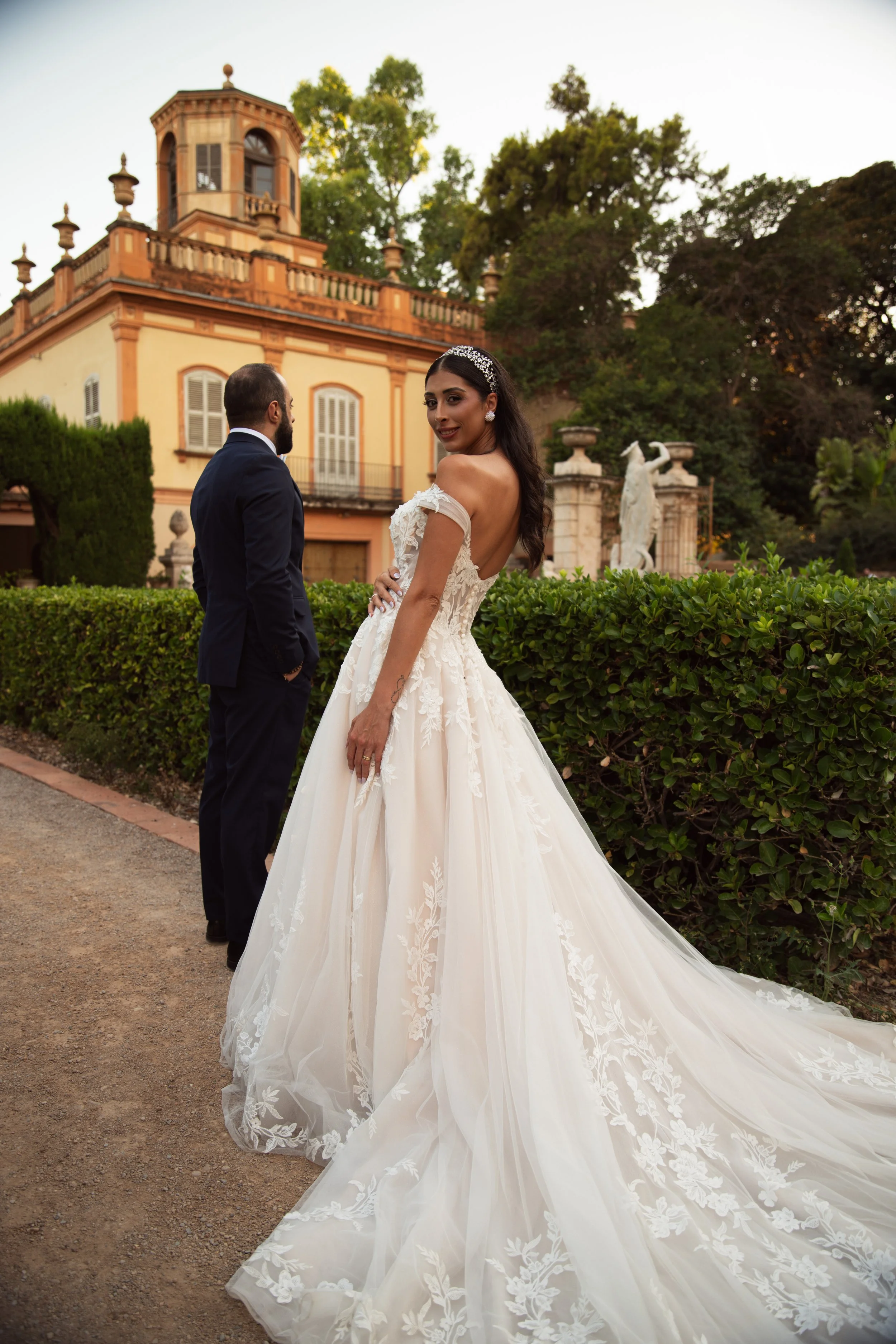 Mujer con vestido de novia y mujer con traje formal en un jardín con historia y arquitectura clásica.