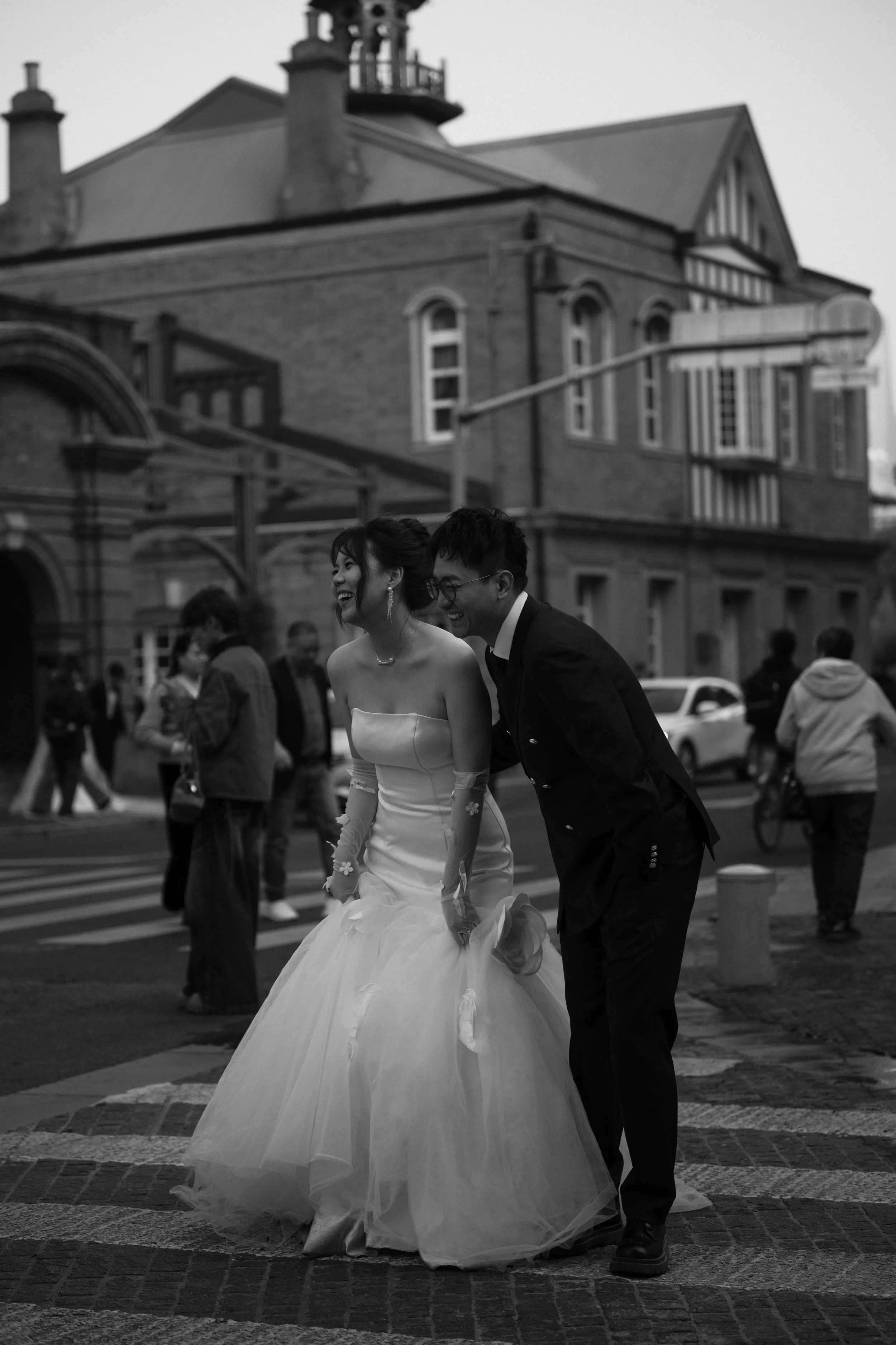 Pareja de novios riendo mientras caminan en la calle, ella con vestido de novia y él con traje, en un día nublado, en un entorno urbano con otras personas y edificios de fondo.