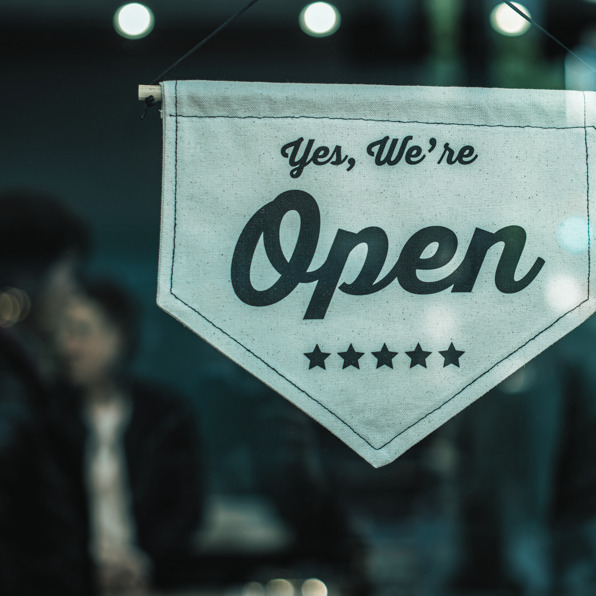 Close-up of a fabric banner with the words "Yes, We're Open" and five stars, hanging with a blurred background of circular lights.