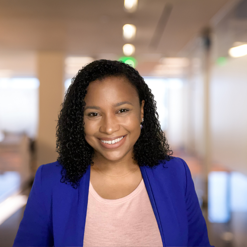A smiling Black woman with curly hair, wearing a blue blazer and a light pink top, standing in a modern office environment.