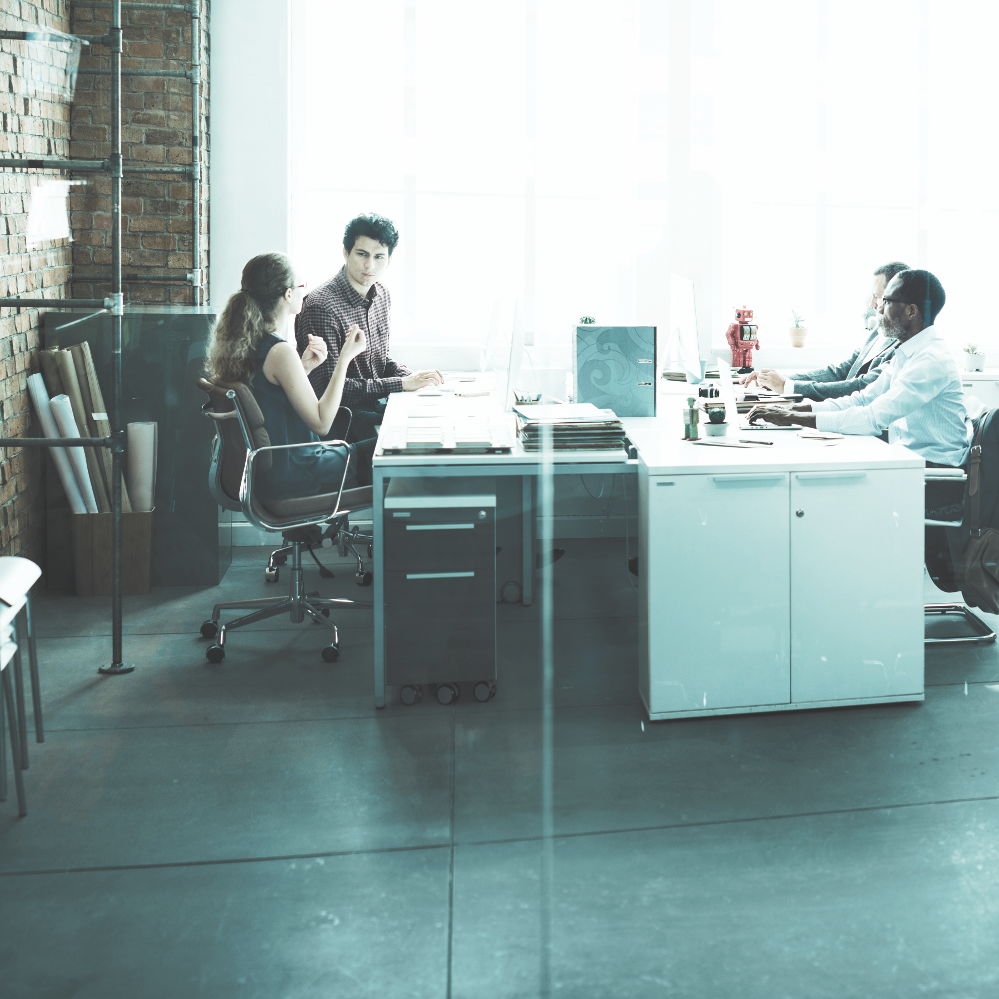 View through a glass door showing a modern office with four people sitting at a long white table, working on computers. The office has large windows, a brick wall on the left, and minimalist decor.