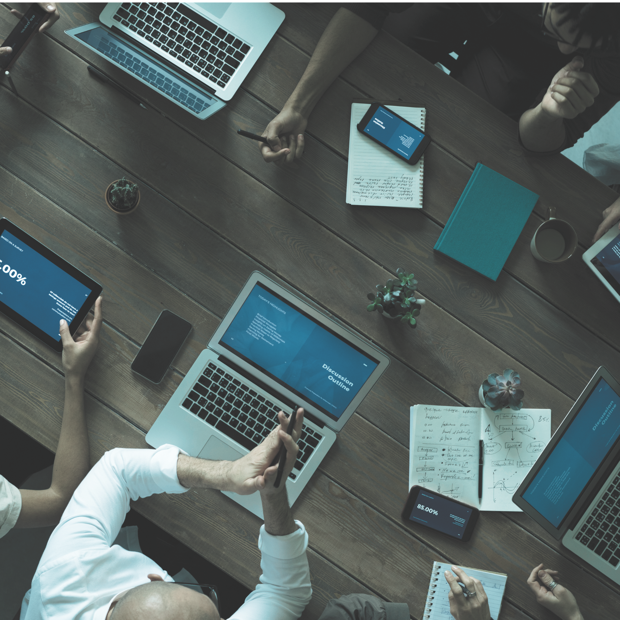 Overhead view of people working at a wooden table with laptops, tablets, notebooks, and smartphones, analyzing data and writing notes.