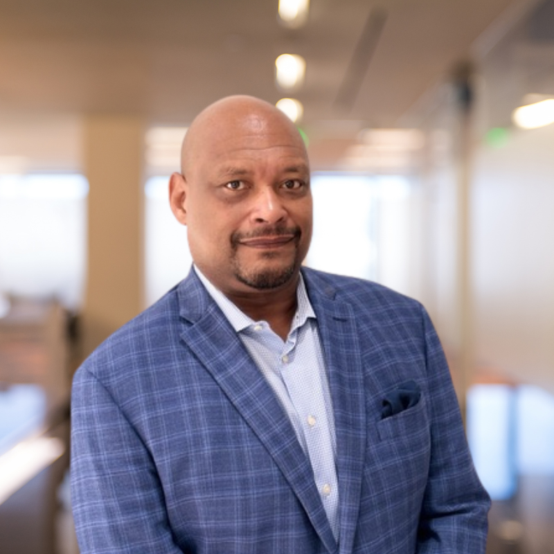 A confident man in a blue plaid blazer stands in a bright, modern office space with blurred background.