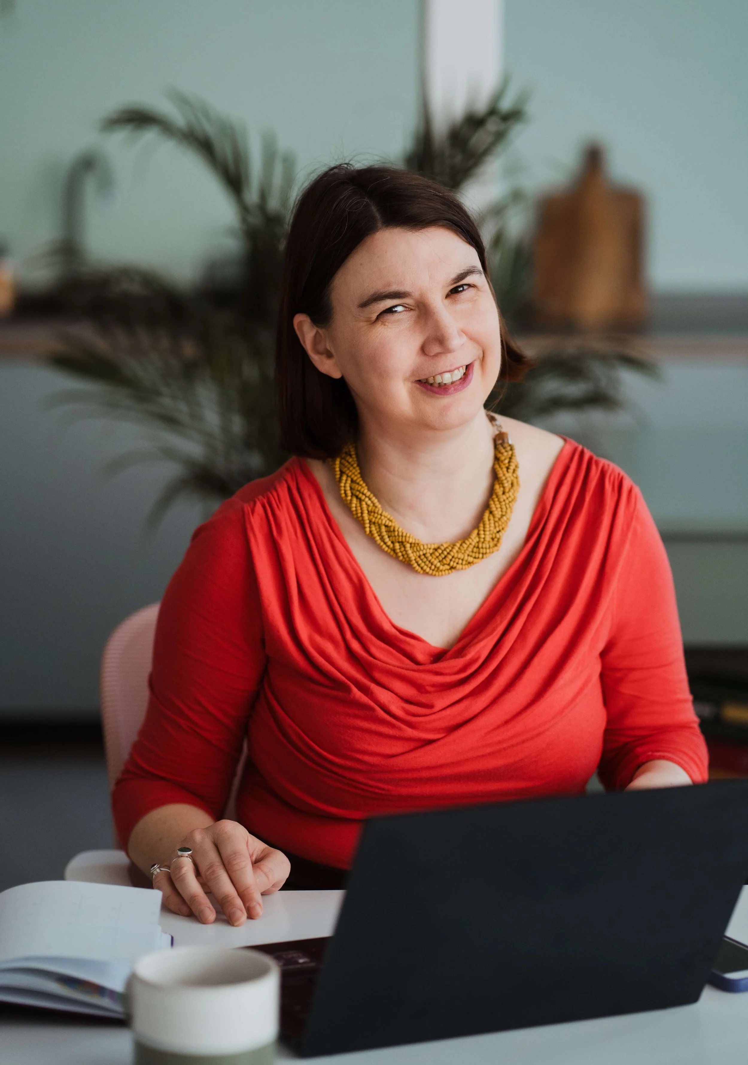 Kathryn is an Disabled People's access and inclusion consultant. She is sat at her laptop, in a red top in front of a plant