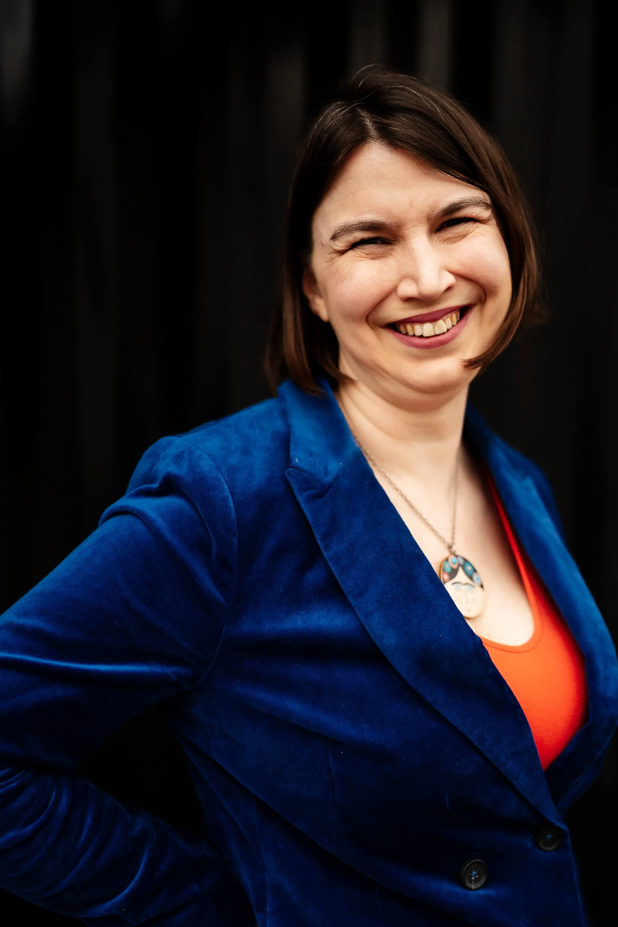 Kathryn, a white woman with brown hair, smiles at the camera. She is an access and inclusion consultant in a blue suit and a Frida Kahlo necklace. She's in front of a dark background