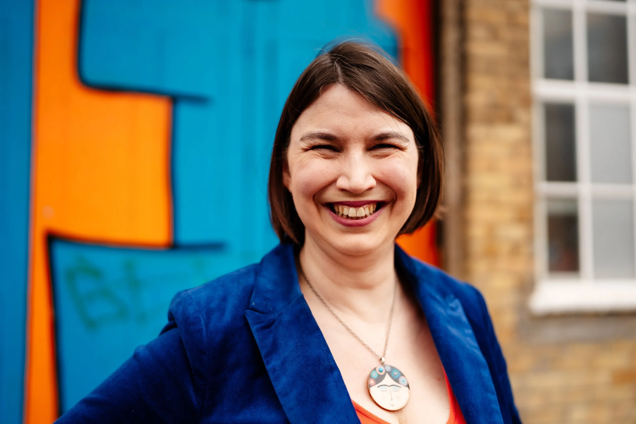 Kathryn, A woman with short brown hair smiling, wearing a blue blazer and a necklace with a Frida Kahlo pendant, in front of a colorful painted wall.