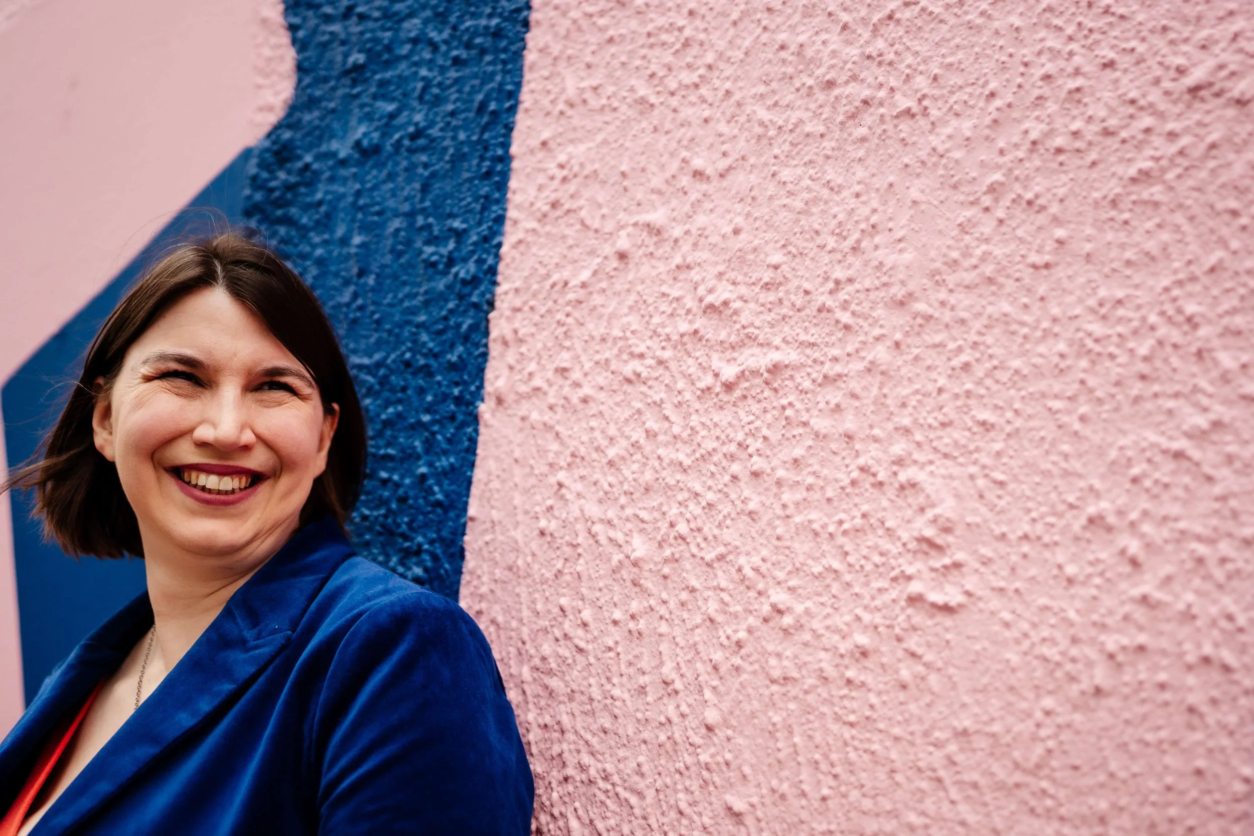 Kathryn, a white woman with brown hair, Looks to her left, in front of a pink and blue backgroundsmil. She is an access and inclusion consultant in a blue suit and a Frida Kahlo necklace.