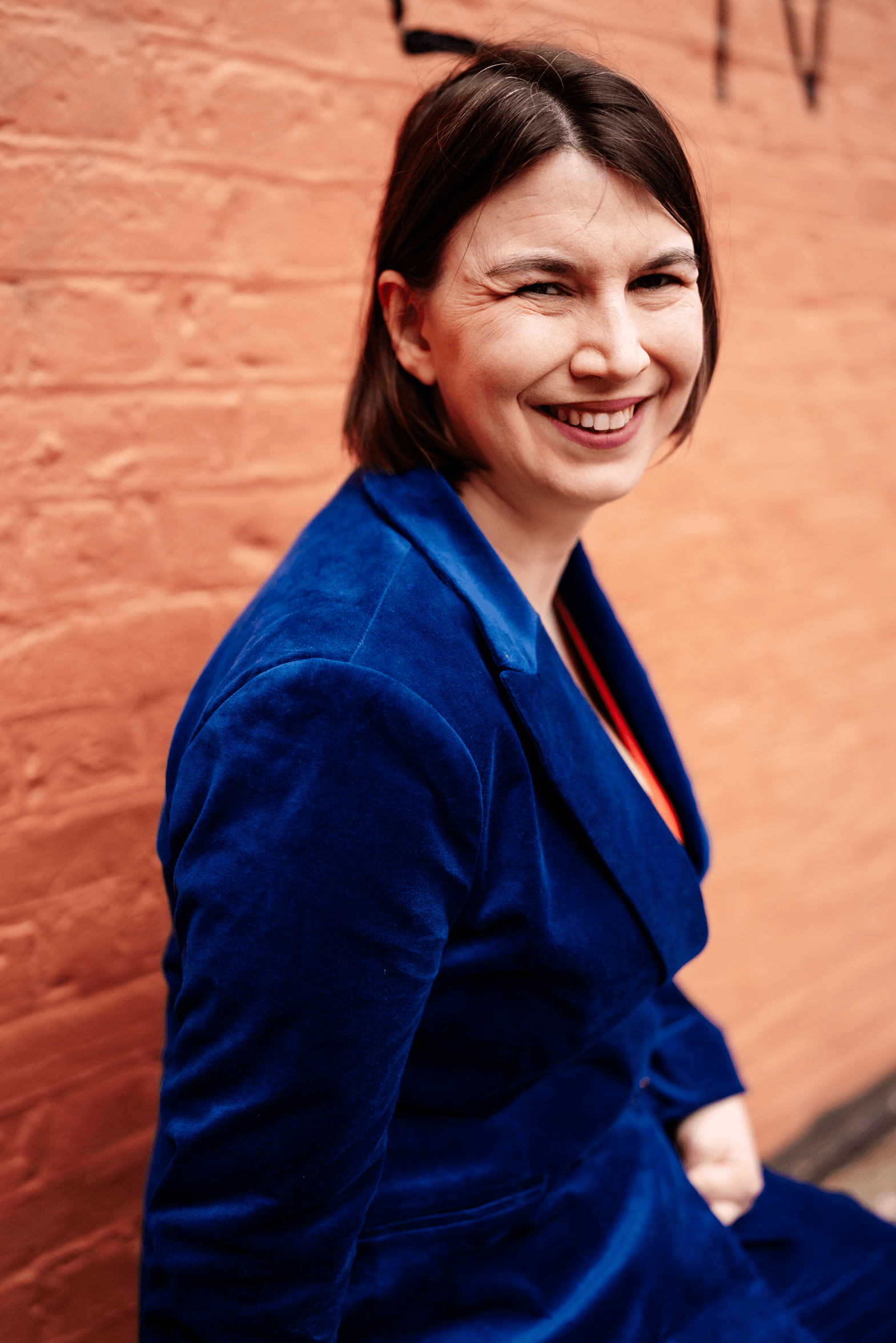 Kathryn, a white woman with brown hair, smiles at the camera over her right shoulder. She is an access and inclusion consultant in a blue suit and a Frida Kahlo necklace.