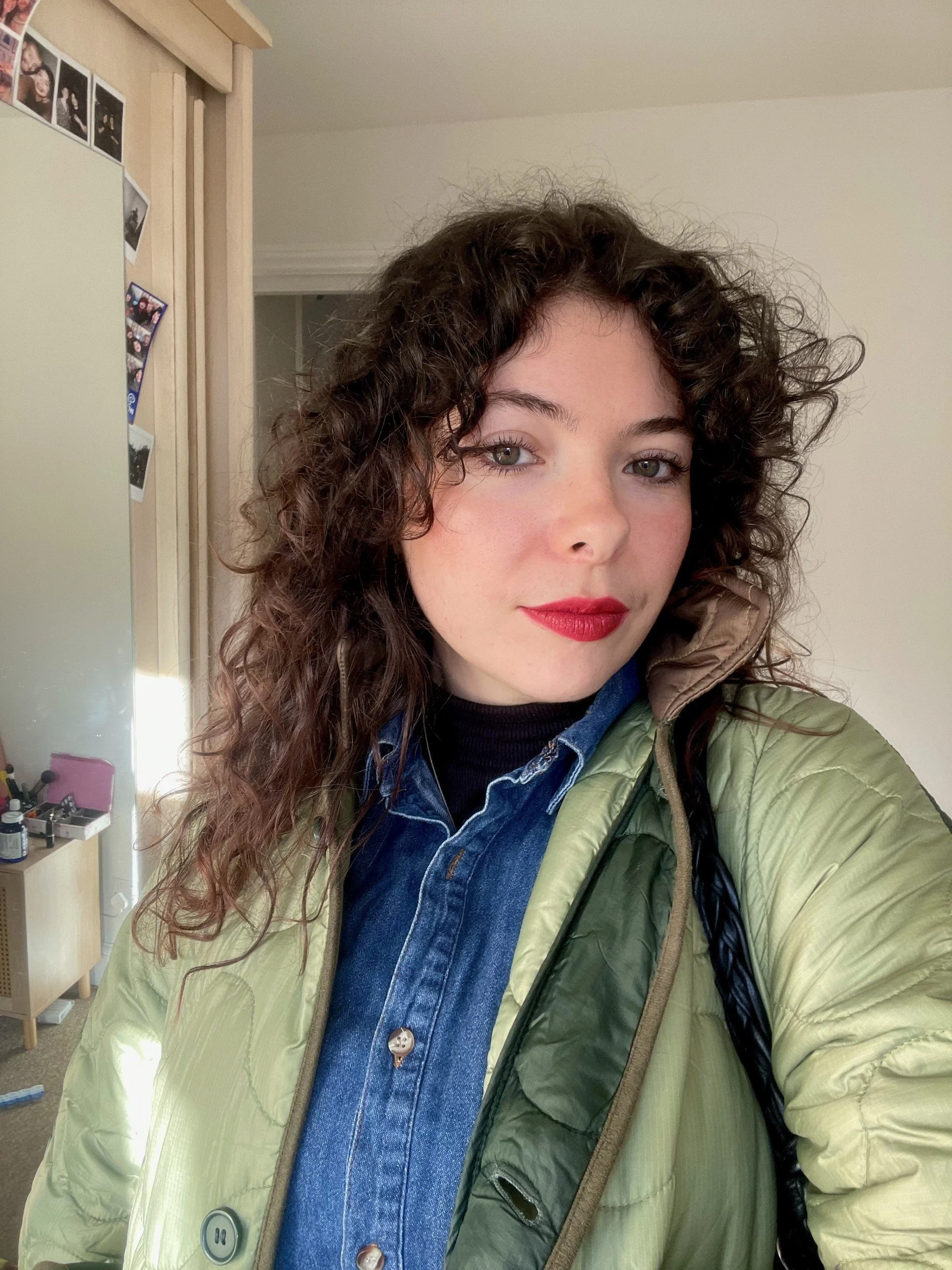 A young woman with curly brown hair taking a selfie indoors, wearing a green puffer jacket over a denim shirt and a black top, with makeup including red lipstick.