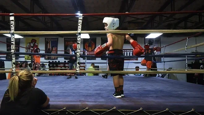 Child in boxing ring wearing a white headgear, red boxing gloves, and athletic shorts, with a coach or trainer in the background.