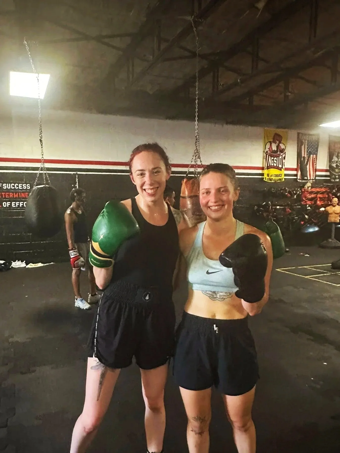 Two women in boxing gear smiling and posing in a gym with boxing equipment and posters in the background.