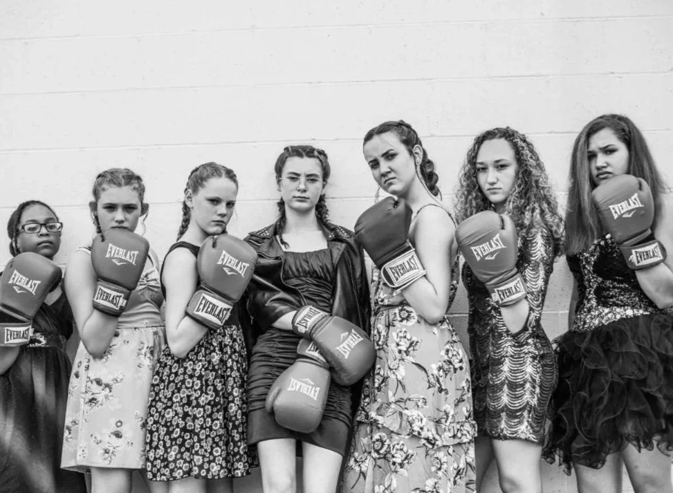 A group of eight young women in boxing gloves, standing against a wall and looking serious.