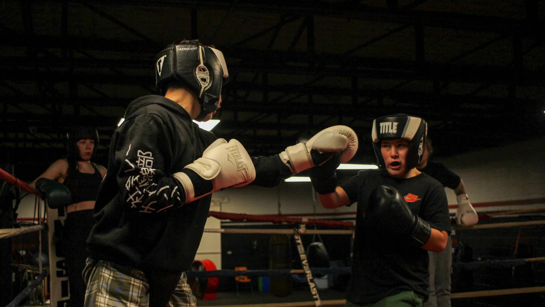 Two people in a boxing gym engaging in a training session, both wearing headgear, gloves, and sportswear, with additional individuals observing in the background.