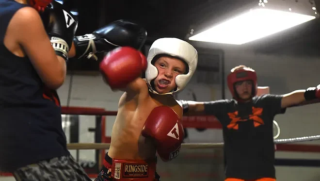 A young boxer in a white helmet and red boxing gloves throws a punch during a sparring match. Another boxer in black gloves and attire is partially visible on the left. A person in a red helmet and black shirt stands in the background in a boxing gym.