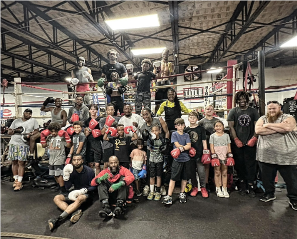 Group of children and adults at a boxing gym, some wearing boxing gloves and standing inside a boxing ring.