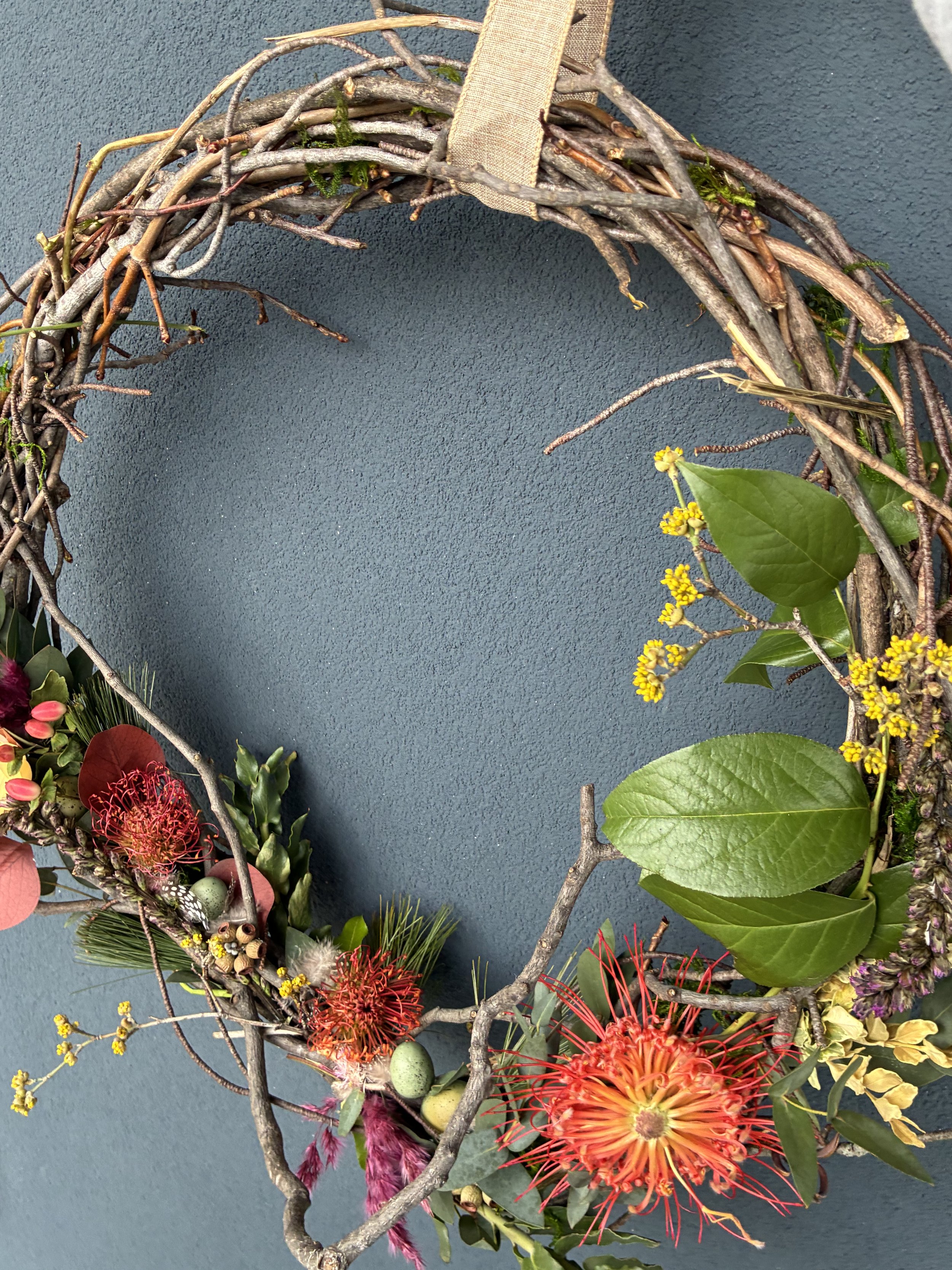 A decorative wreath made of intertwined twigs and branches decorated with bright red and orange flowers, green leaves, and small yellow flowers, hanging against a dark blue wall.