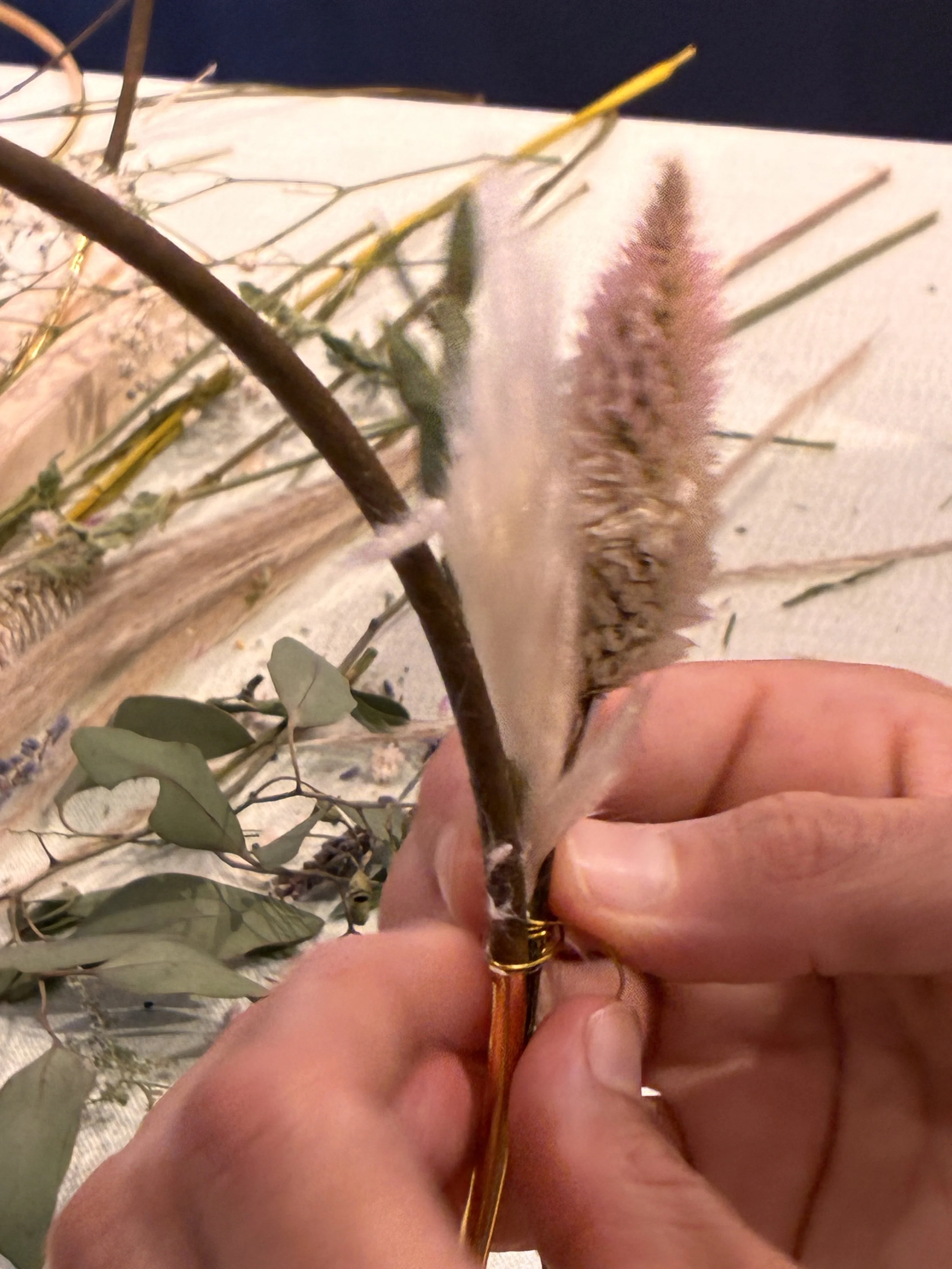 A human hand holding a small branch with eucalyptus and pink celosia flower creating a wreath on a floral hoop.