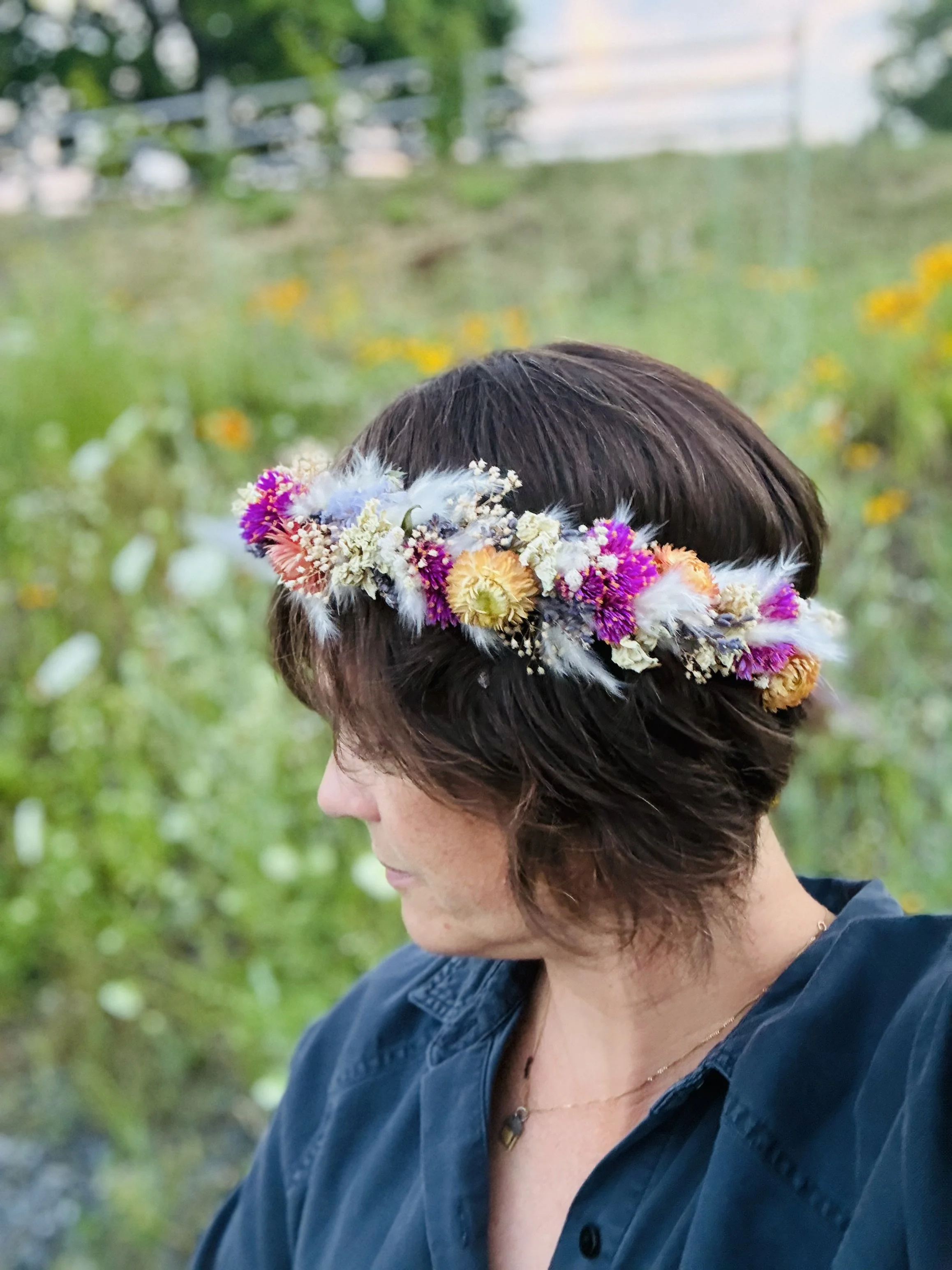 A woman with short brown hair wearing a floral crown made of colorful flowers and white bunny tail grasses, standing outdoors with blurred greenery in the background.