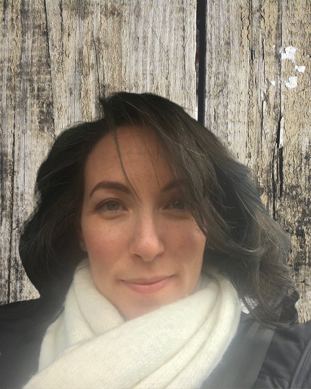 A woman with dark, wavy hair and fair skin poses in front of a weathered wooden wall, wearing a white scarf and a dark jacket.