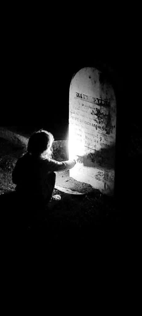 A child sitting at night, illuminated by a small flashlight, reaching for an imposing gravestone.