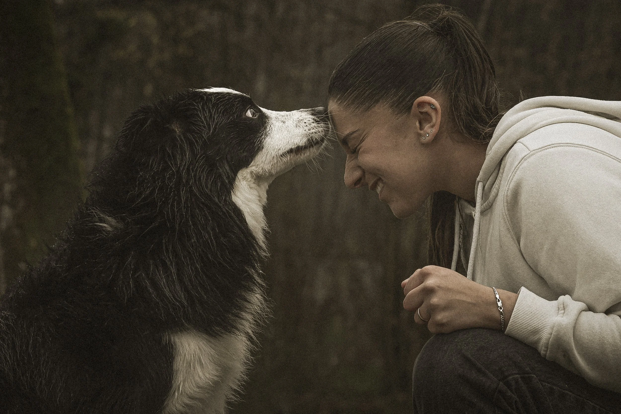 Une jeune femme, de profil, sourit avec ses yeux fermés, face à un chien de race Border Collie noir et blanc. La femme porte une veste blanche à capuche et a plusieurs piercings à l'oreille. Le chien regarde la femme et semble calme et attentif. L'ar